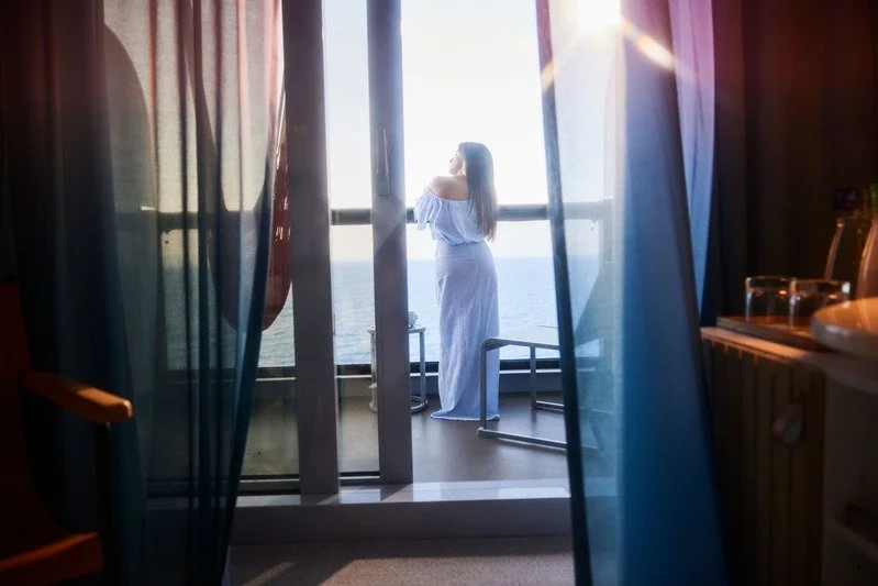 A woman standing on a balcony overlooking the ocean, seen through sliding glass doors with curtains.