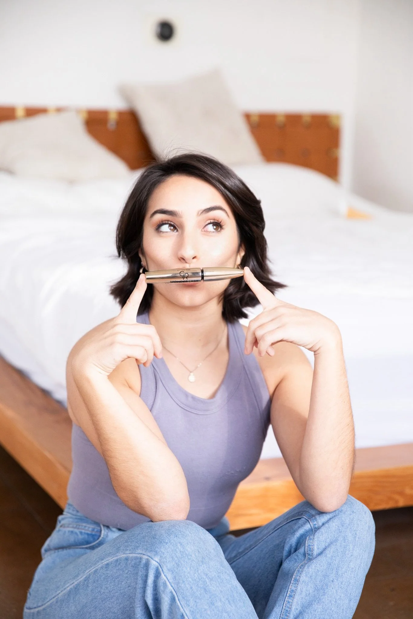 A woman sitting on the floor next to a bed holding a pen horizontally across her face, with her fingers pointing to her cheeks and looking upward.