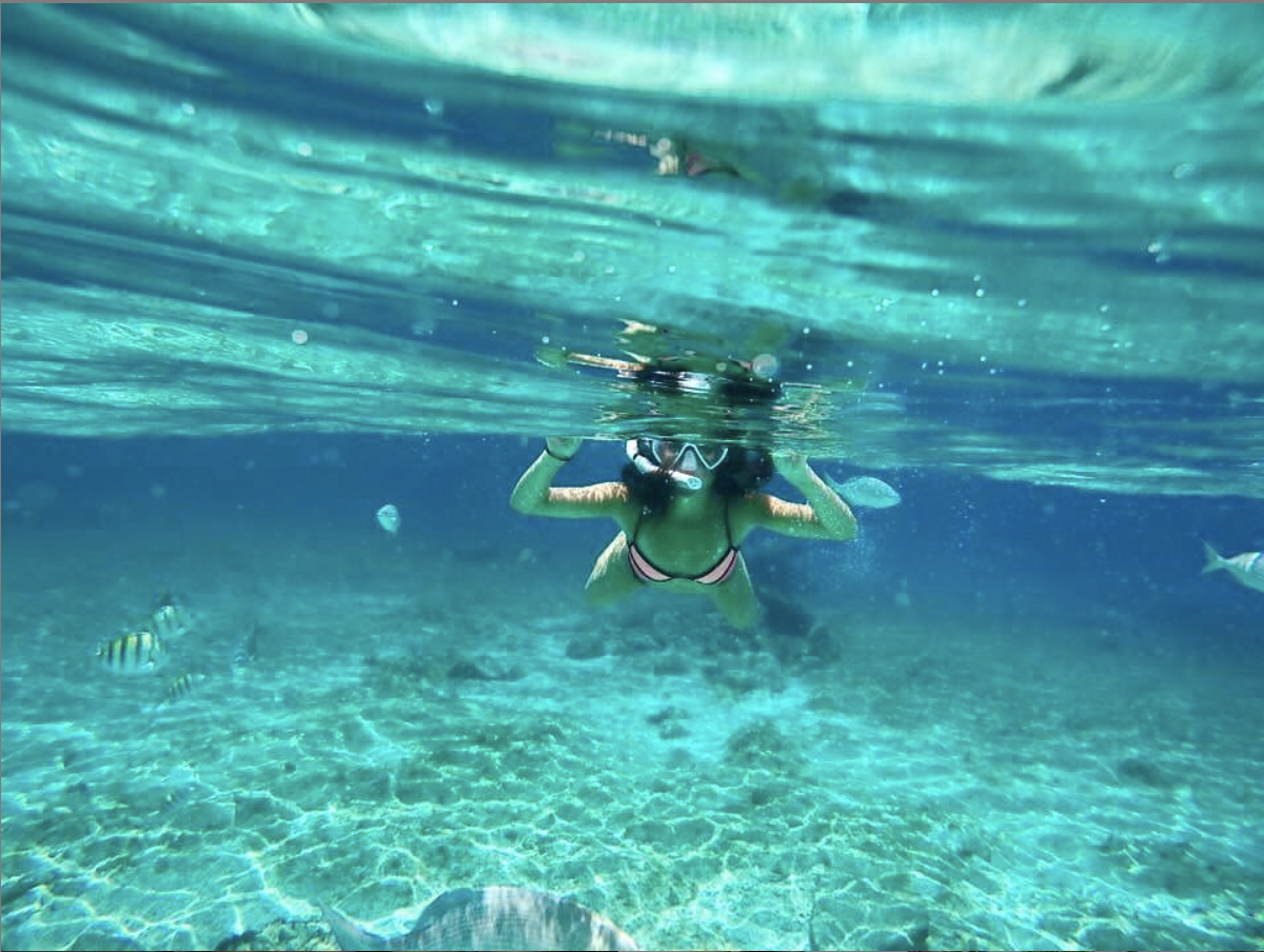 A person snorkeling underwater, wearing a mask and fins, surrounded by fish in clear blue water.