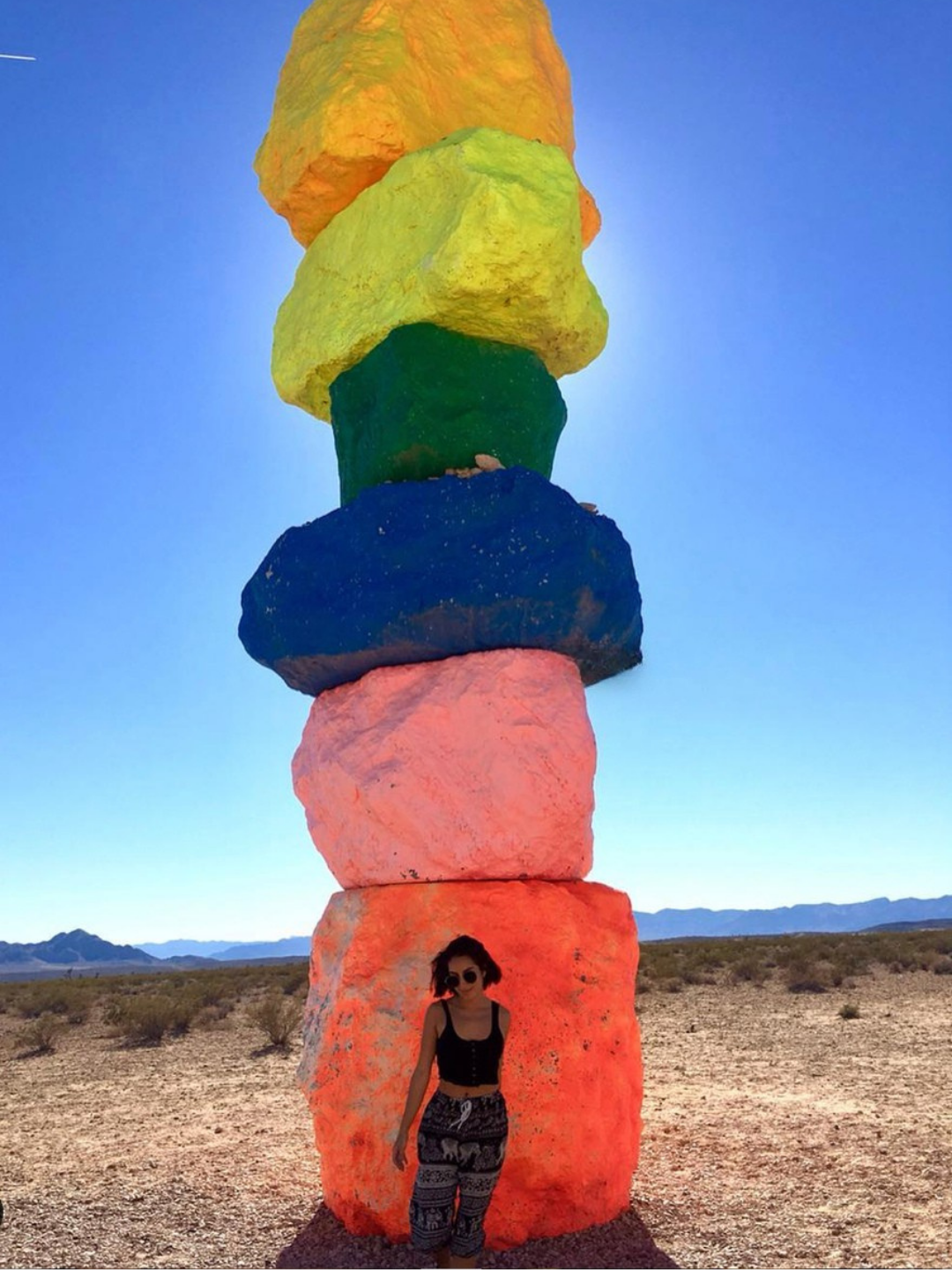 A woman stands in front of a stack of large, brightly colored rocks in a desert landscape, with mountains in the background and a clear blue sky overhead.