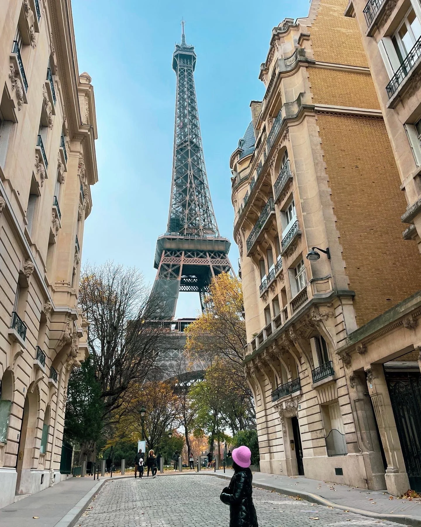 Looking up at the Eiffel Tower from a Paris street with classic Parisian buildings on either side, a woman in a pink hat and black coat stands in the foreground, with trees and a few pedestrians in the background.