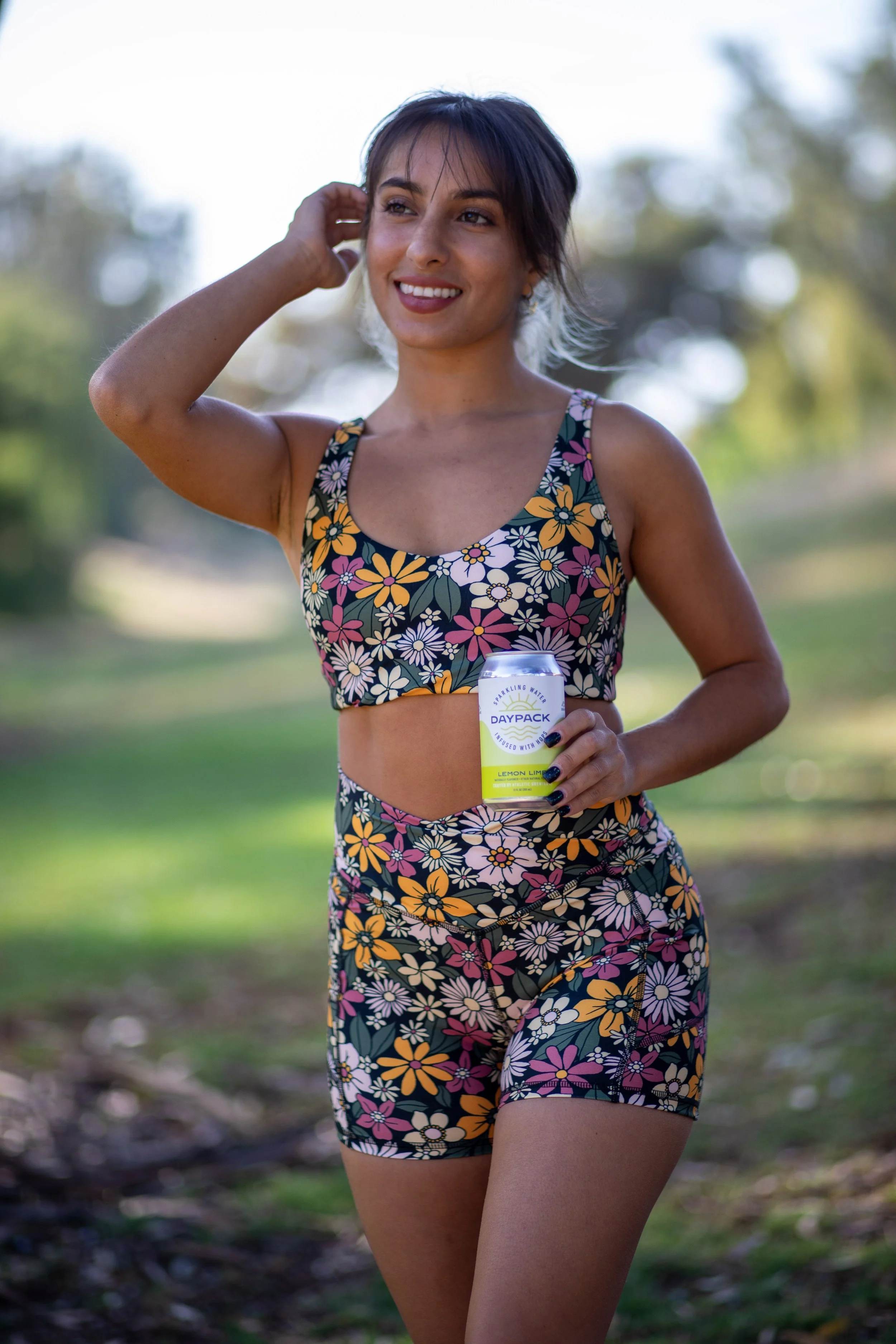 Young woman in floral sportswear holding a can of Lemon Lime flavored sparkling water, standing outdoors on a sunny day.