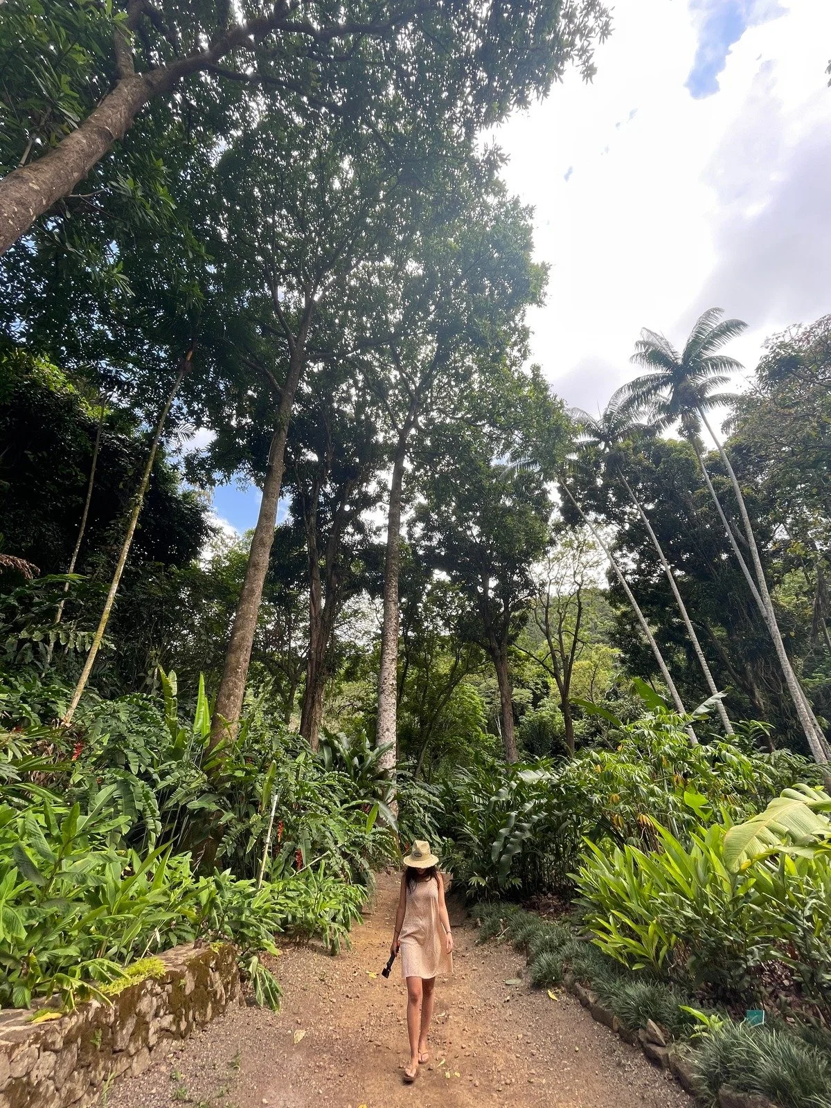 A woman in a beige dress and a wide-brimmed hat walking along a dirt path surrounded by tall, lush green tropical trees and plants.