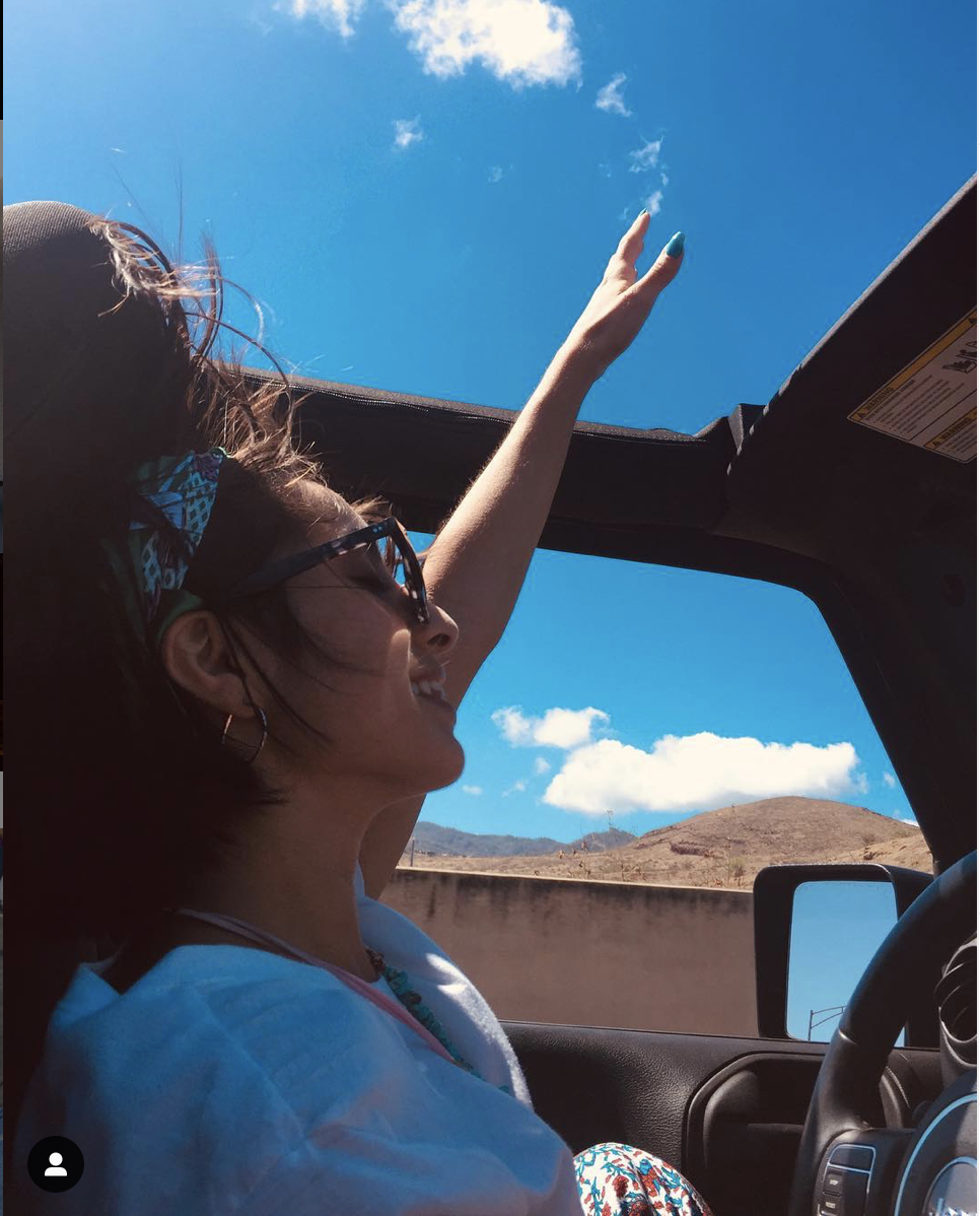 Woman with short hair wearing glasses, sitting in a convertible car, reaching up towards the sky with her right hand, smiling, blue sky with clouds and distant hills in the background.