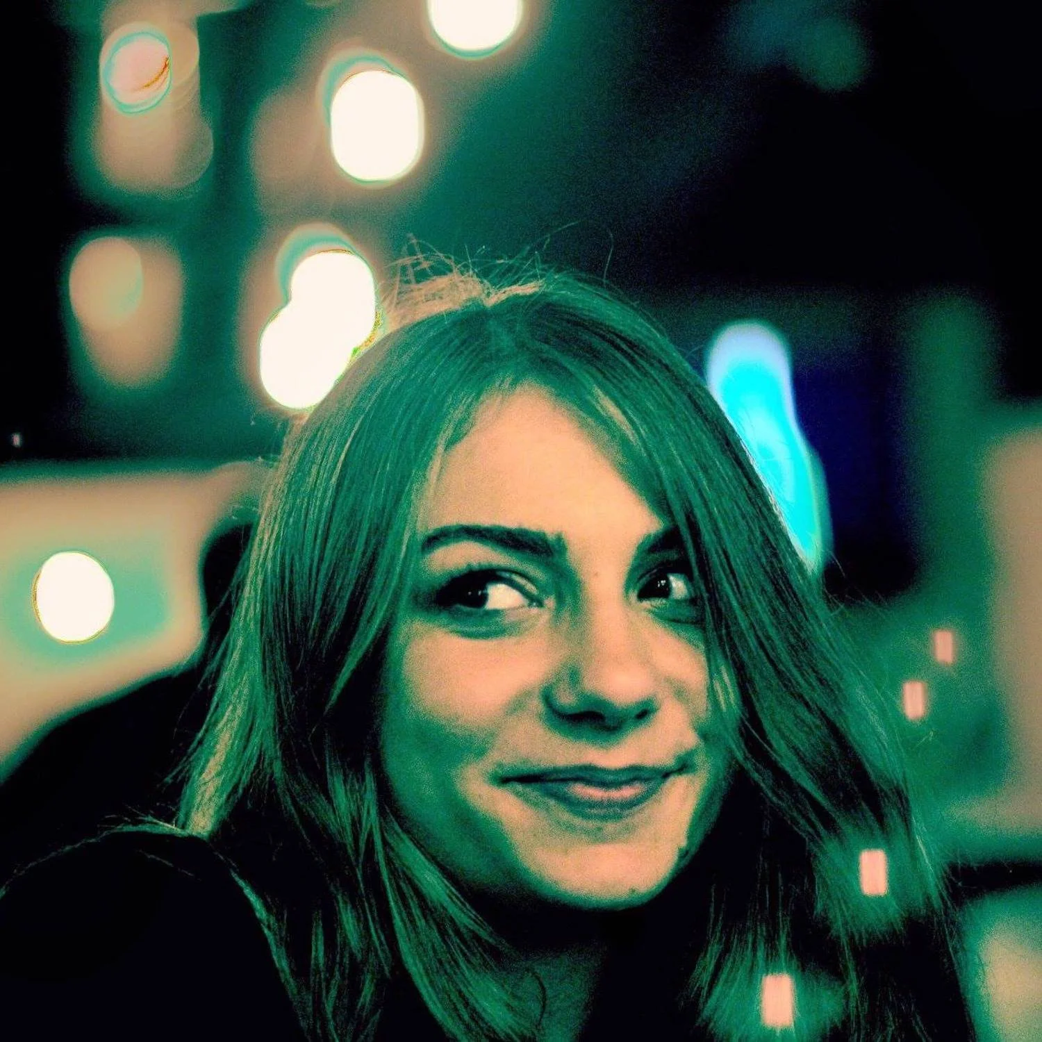 A woman with long hair smiling at night, with colorful bokeh lights in the background.