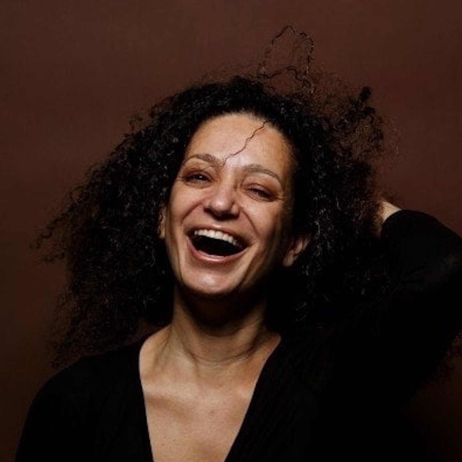 A woman with curly black hair laughing and posing with one hand behind her head against a brown background.