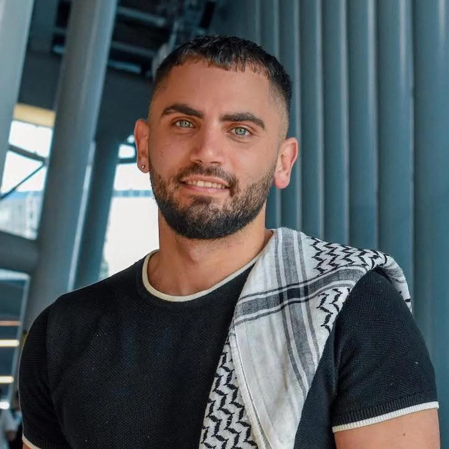 Portrait of a young man with short dark hair and a beard, wearing a black shirt, posing in front of a modern architectural background with blue vertical panels and structural elements.