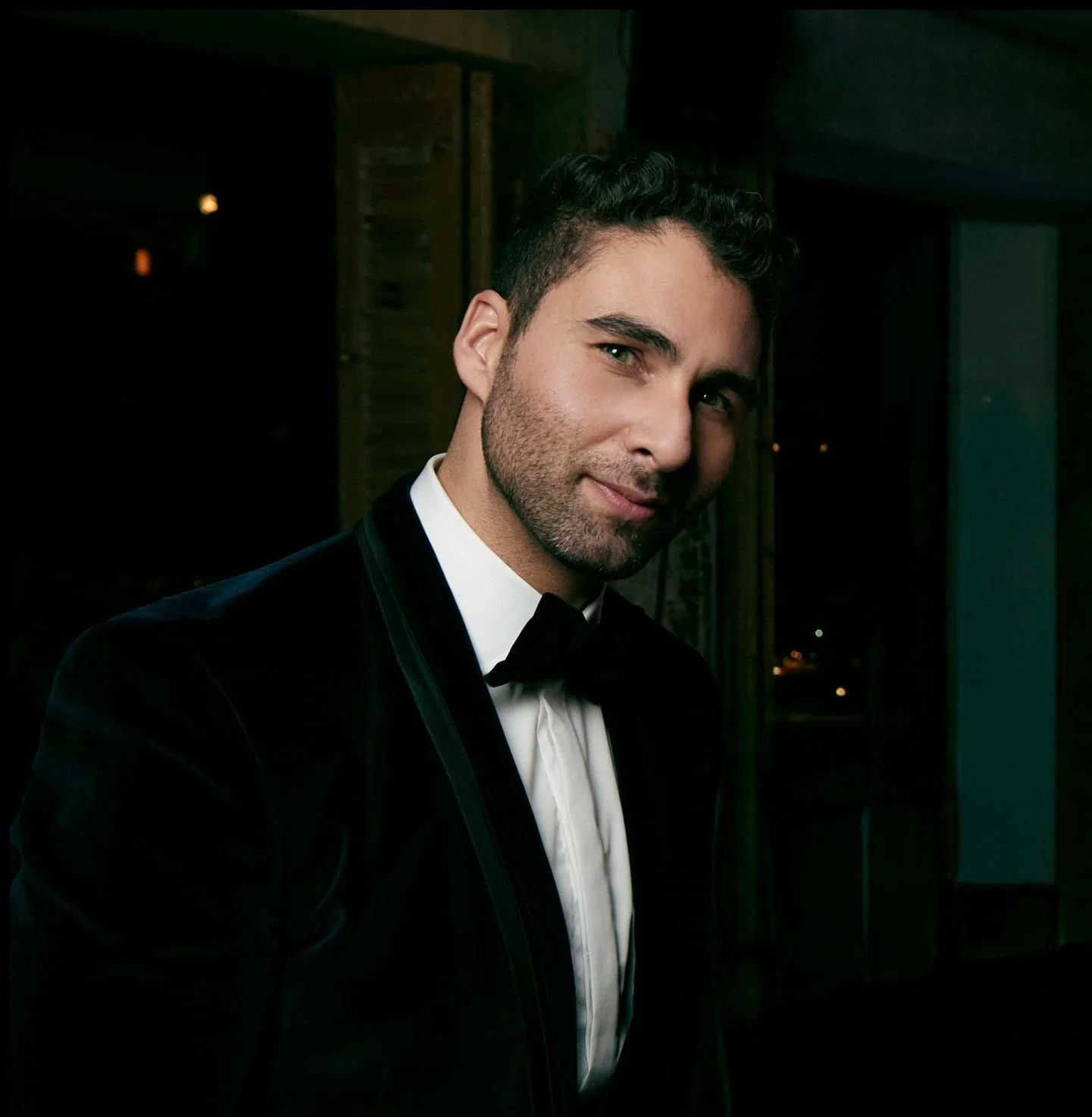 A man in a tuxedo smiling at the camera, evening setting.