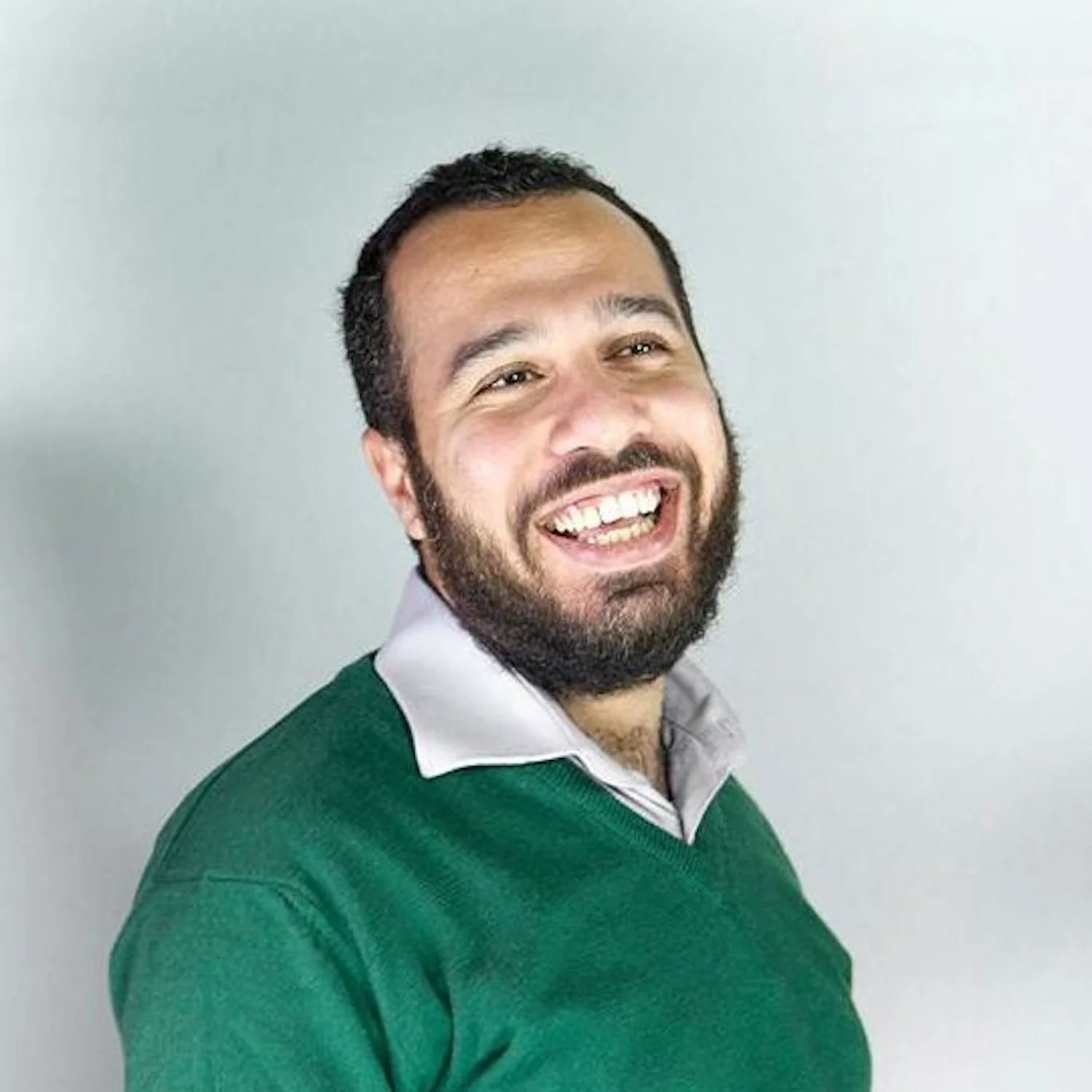 A man with a beard and dark hair, smiling and wearing a green sweater over a collared shirt, standing against a light-colored background.