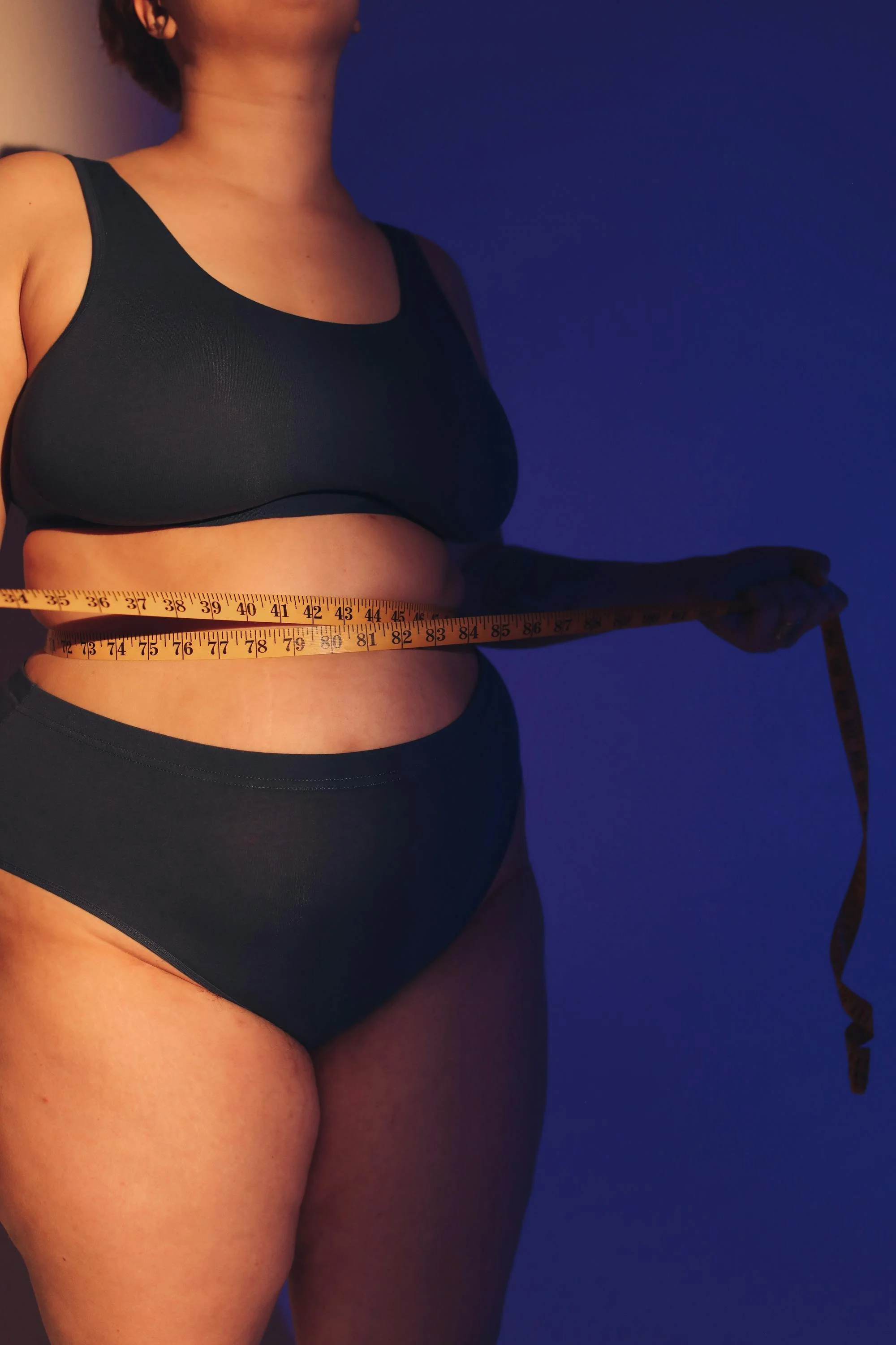 A woman in black underwear measuring her waist with a tape measure in front of a dark blue background.