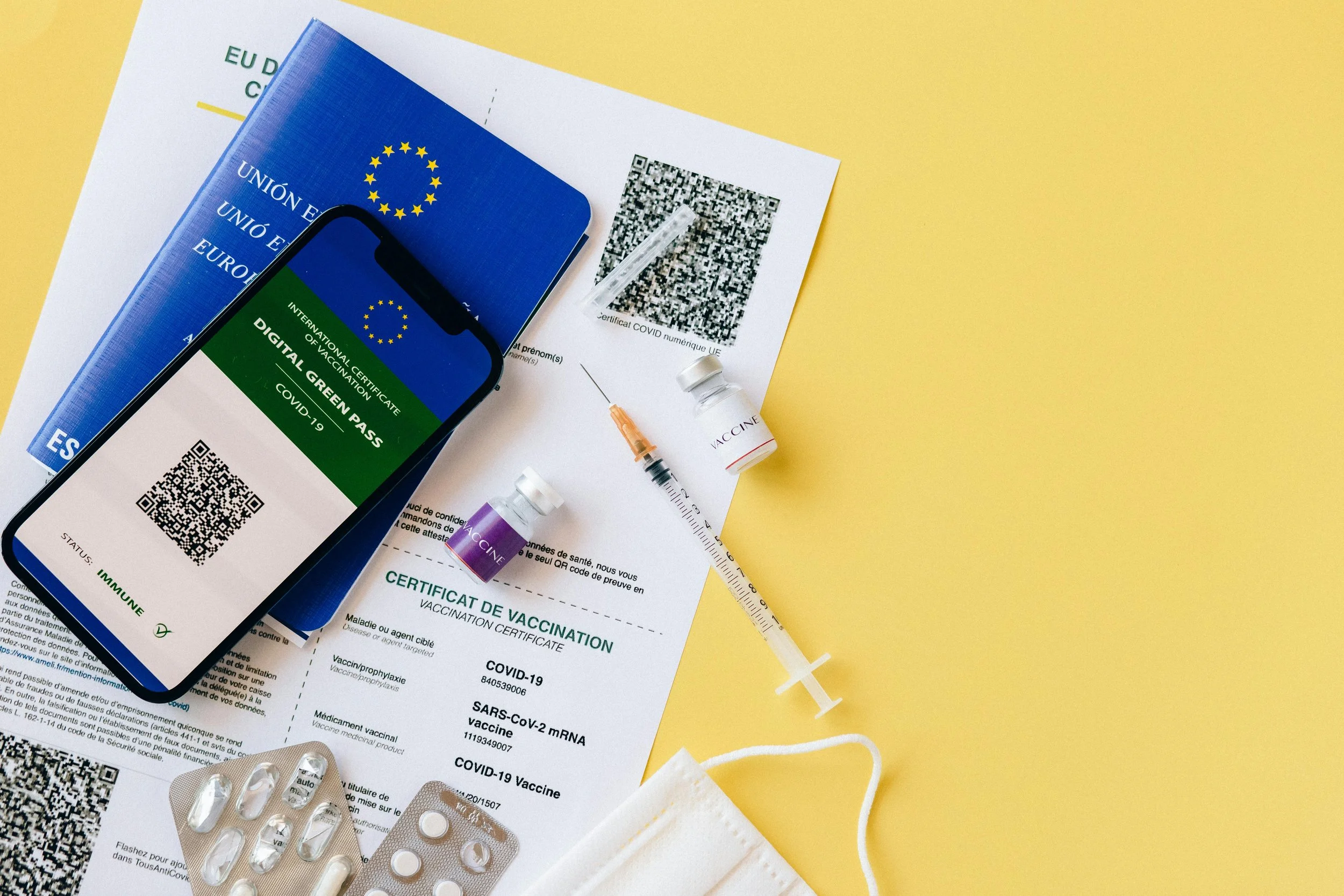 COVID-19 vaccination documents, a mobile phone displaying a digital green pass, vaccines, a syringe, and medication on a yellow background.