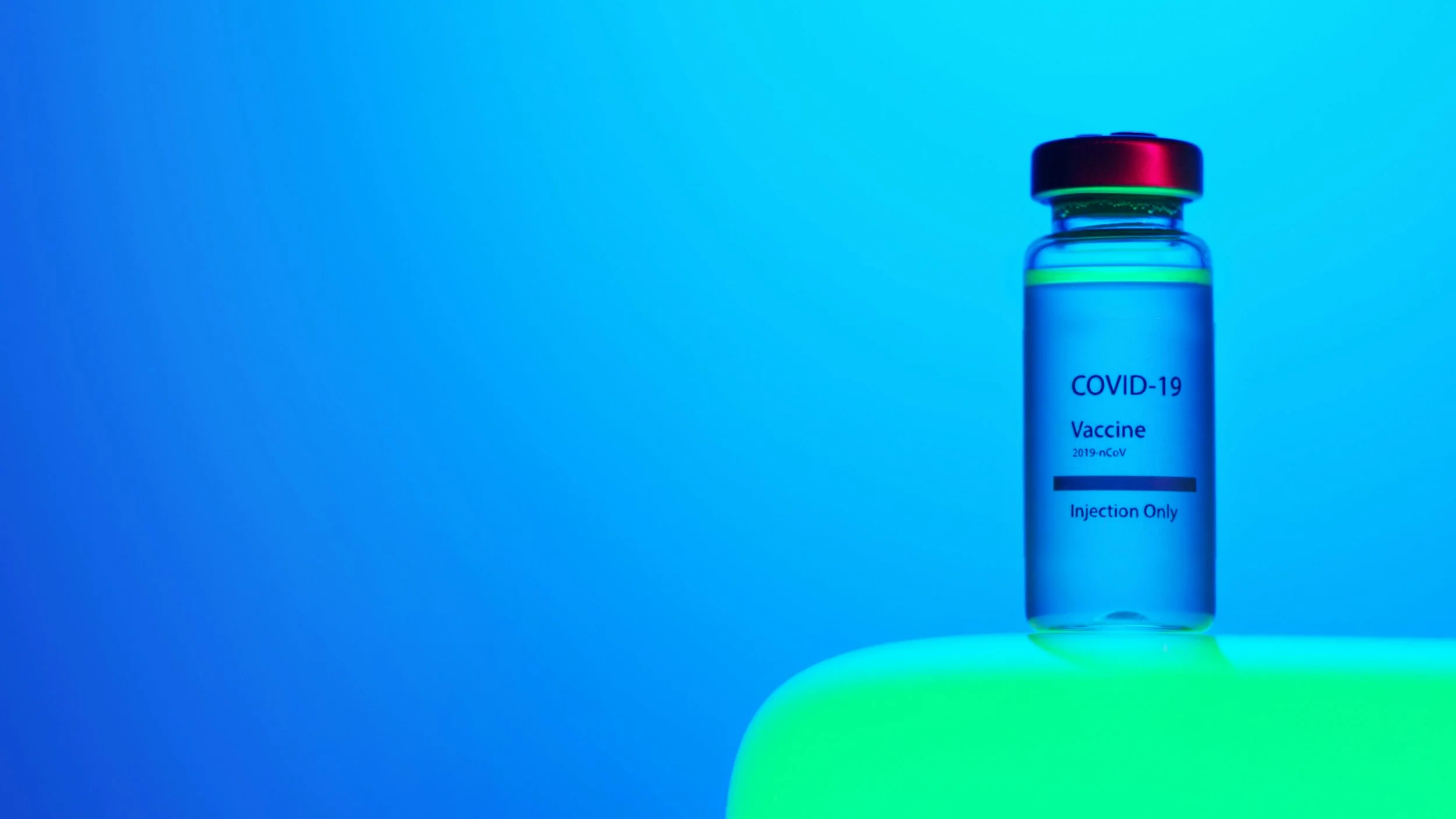 A vial labeled COVID-19 Vaccine with a red cap, illuminated by blue and green lighting, placed on a glowing green surface.