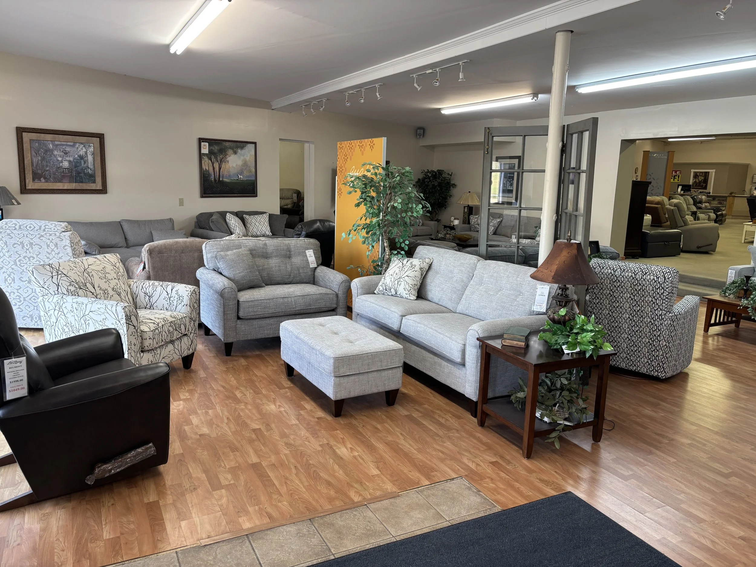 A showroom with various sofas and chairs on a wood floor, with lamps, plants, and framed artwork on the walls.