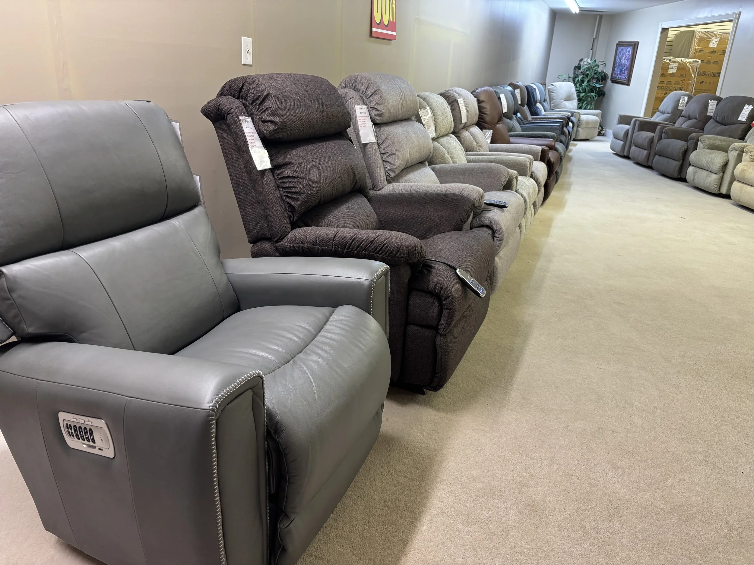 A showroom display of various recliner chairs, arranged in two rows along the walls, with tags attached to each chair.