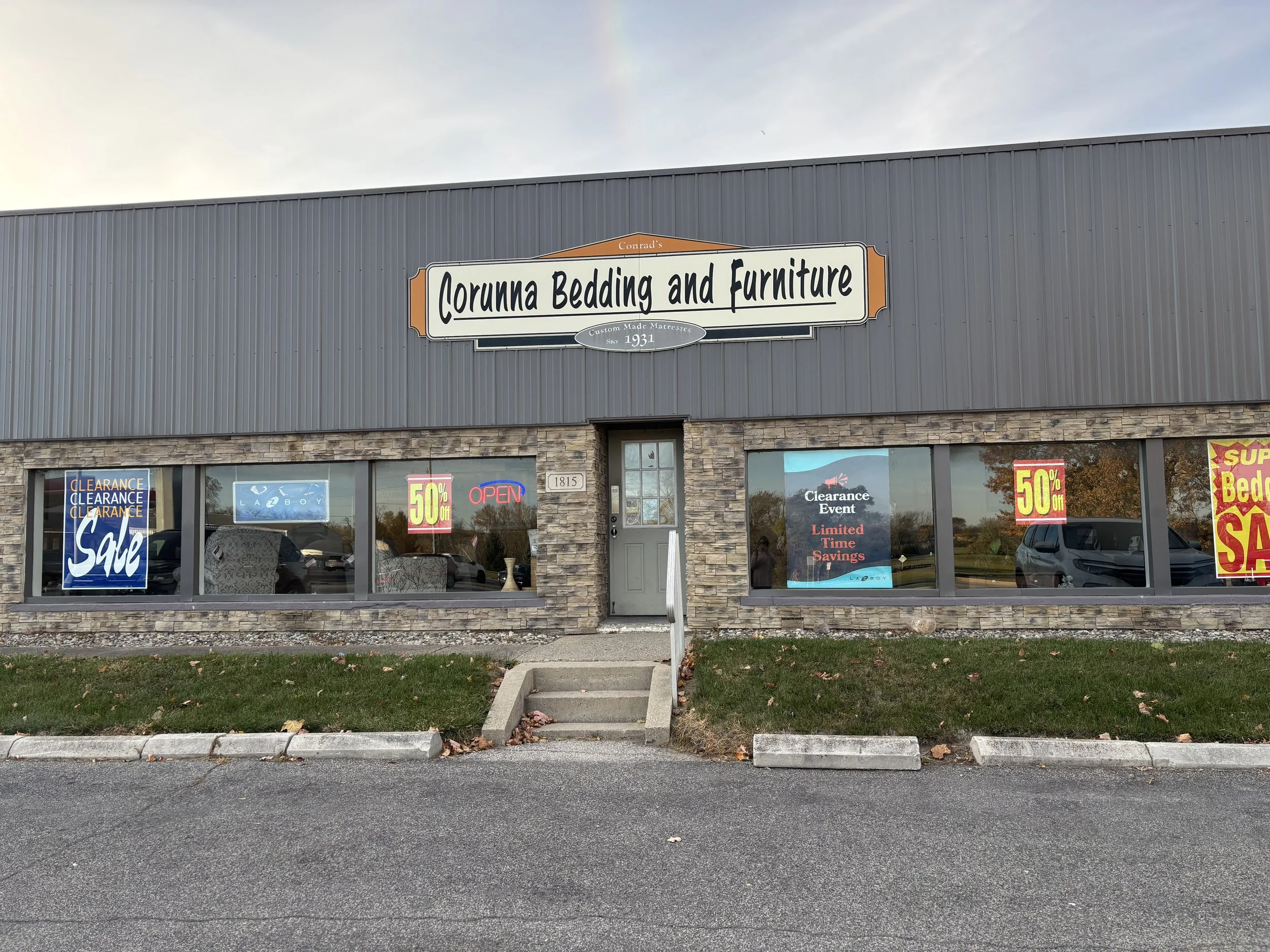 Exterior of a store named 'Corunna Bedding and Furniture' with large windows displaying sale signs and an entrance with a small set of concrete steps.
