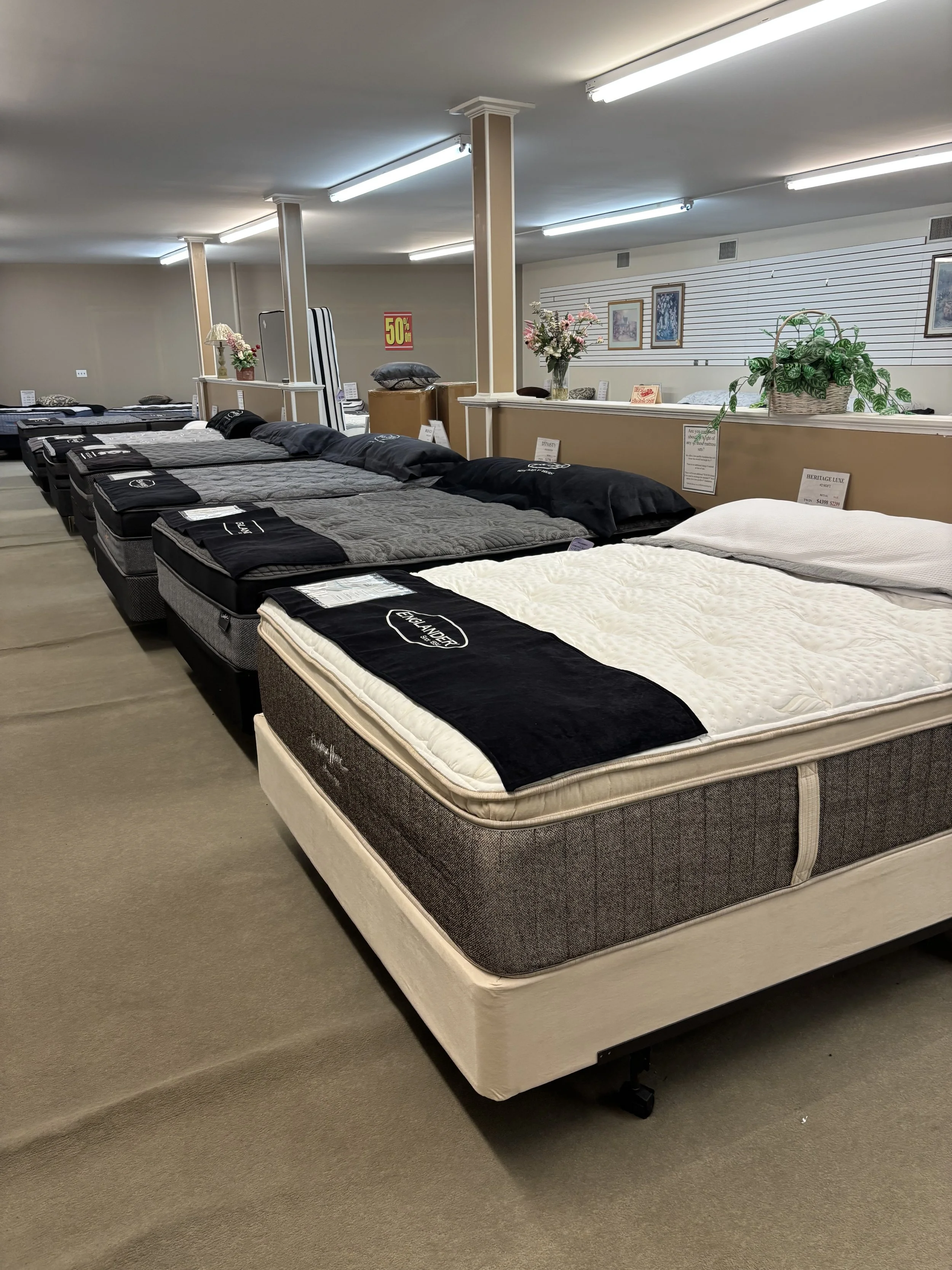 View of a mattress store display with multiple beds lined up, some labeled with tags, several with black, white, and gray bedspreads, and a beige carpeted floor.
