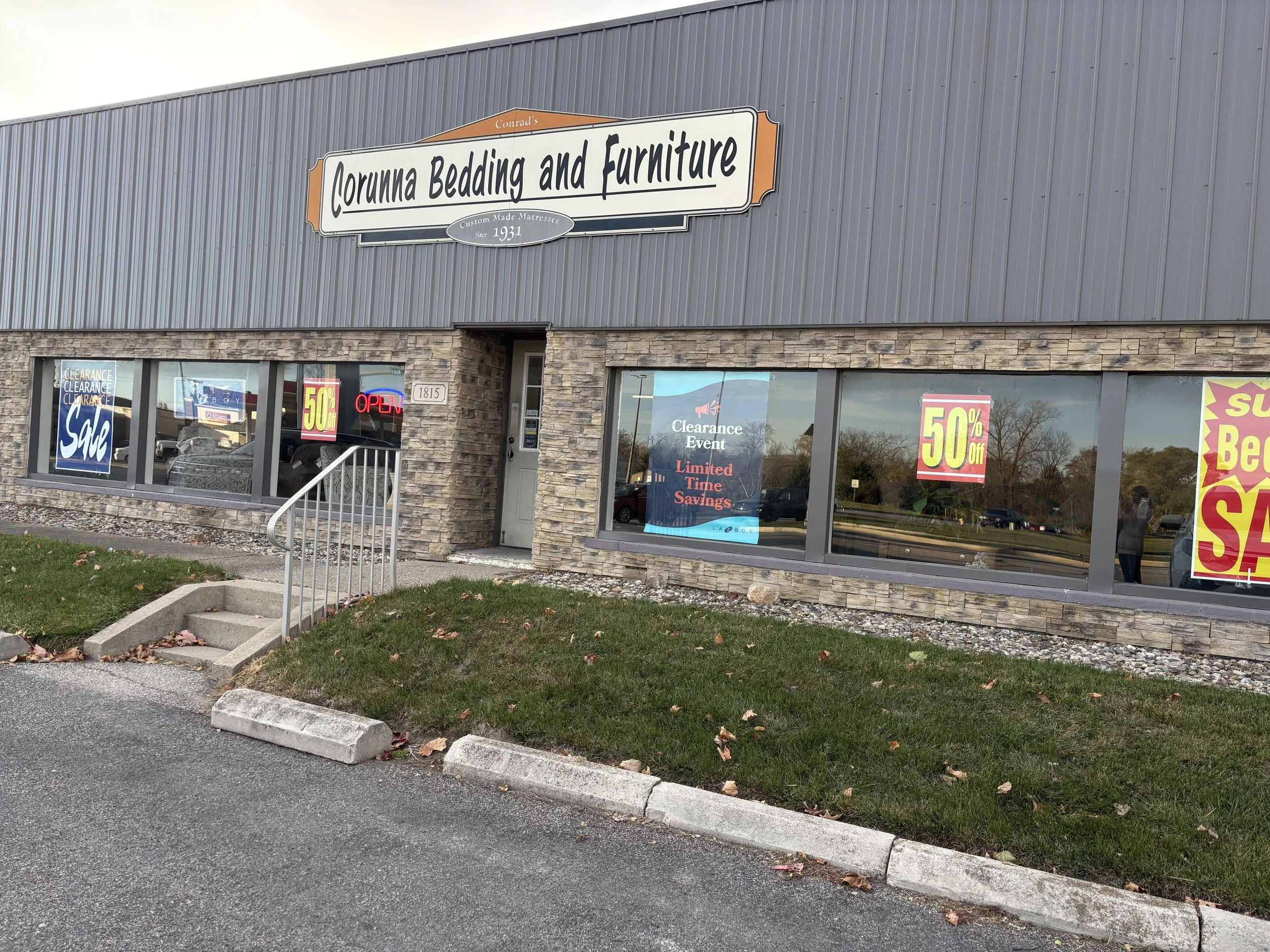 Exterior view of Corunna Bedding and Furniture store with promotional signs in the windows advertising 50% off sale and clearance event, a small set of stairs leading to the entrance, and a grassy area with fallen leaves in front.