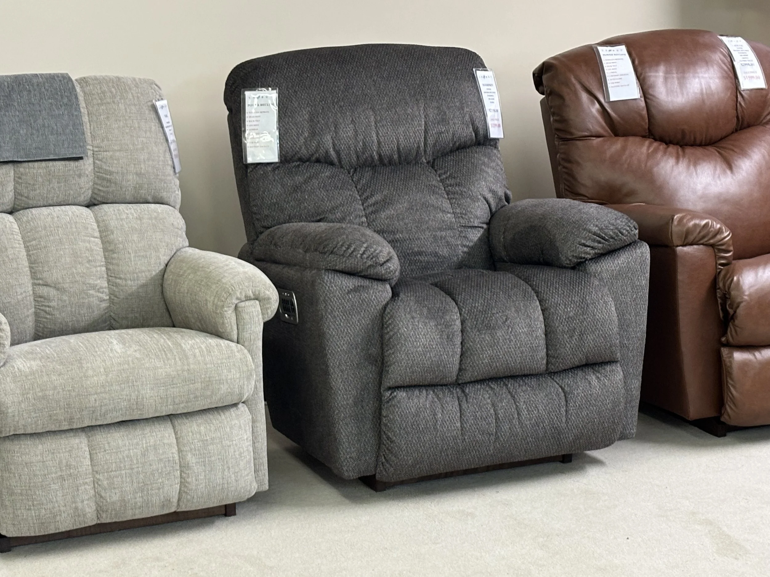 Three recliner chairs in different colors and fabrics are displayed side by side in a furniture store. The left chair is light gray, the middle one dark gray, and the right one brown leather.