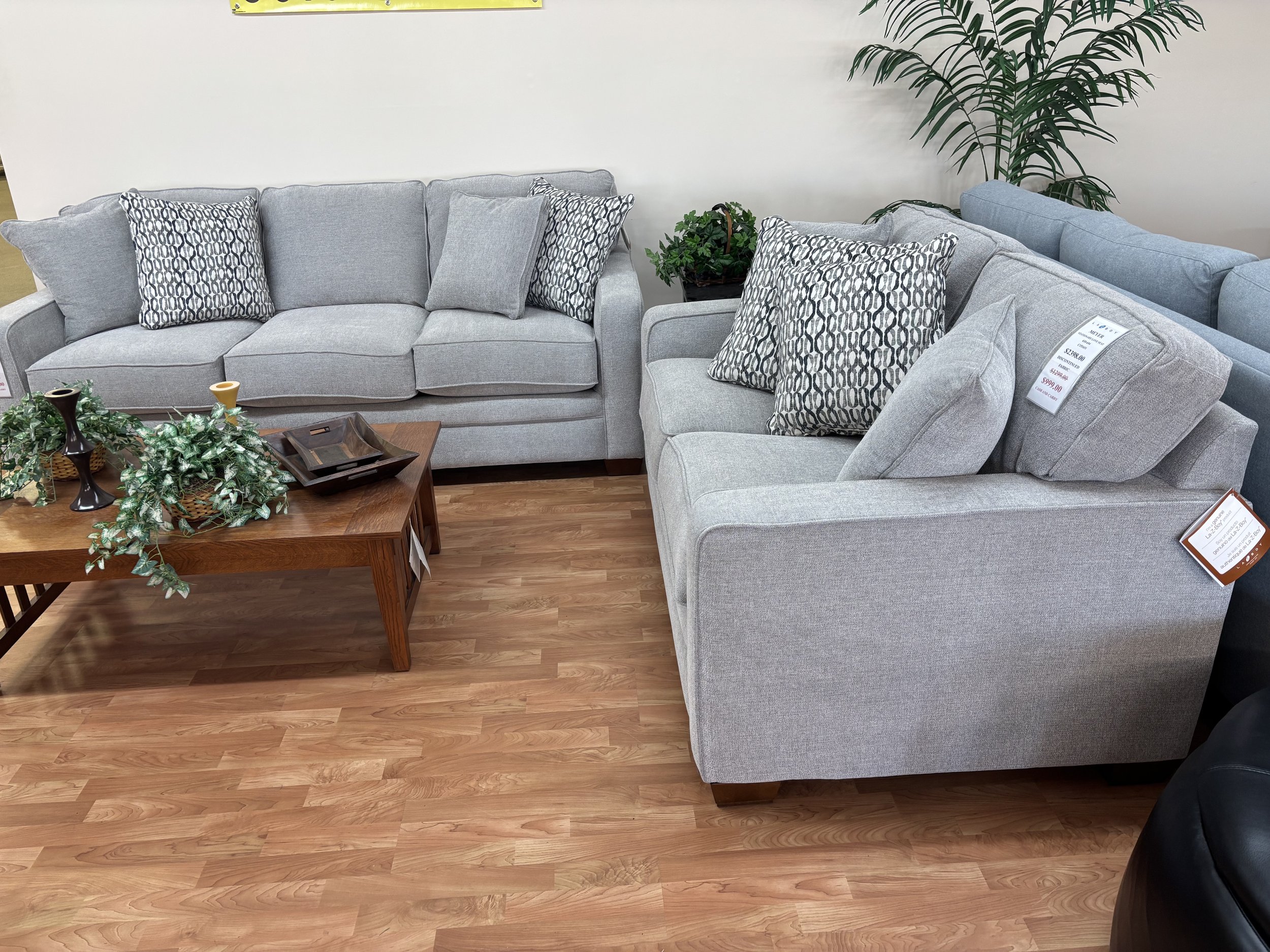Set of four light gray sofas with patterned pillows in a furniture store. Small wooden coffee table with decorative greenery and candle holders in front. A large potted plant and a price tag on the sofa are visible.