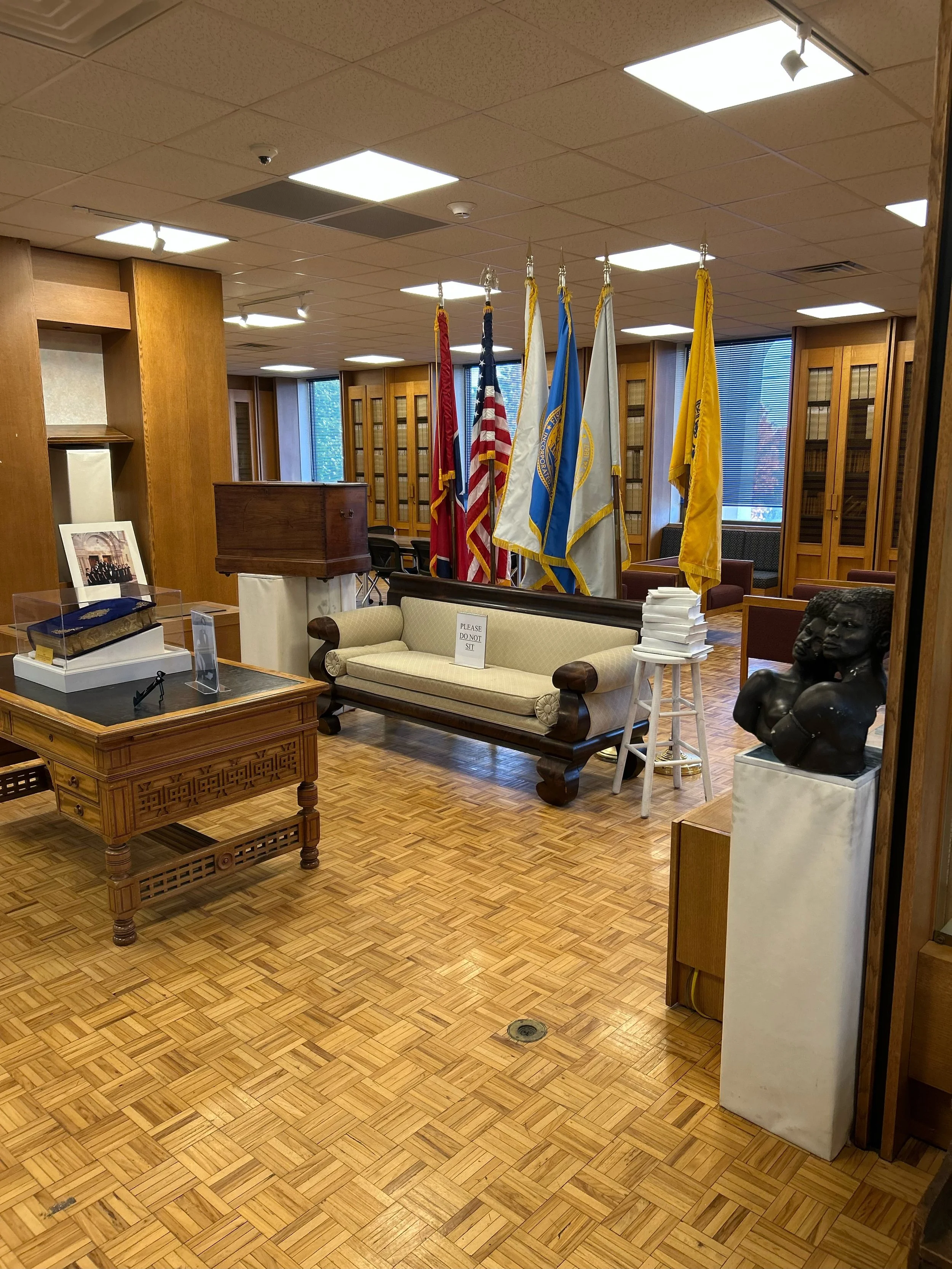 Interior of a room with American flags, a beige sofa, wooden furniture, and a bust sculpture on a pedestal.