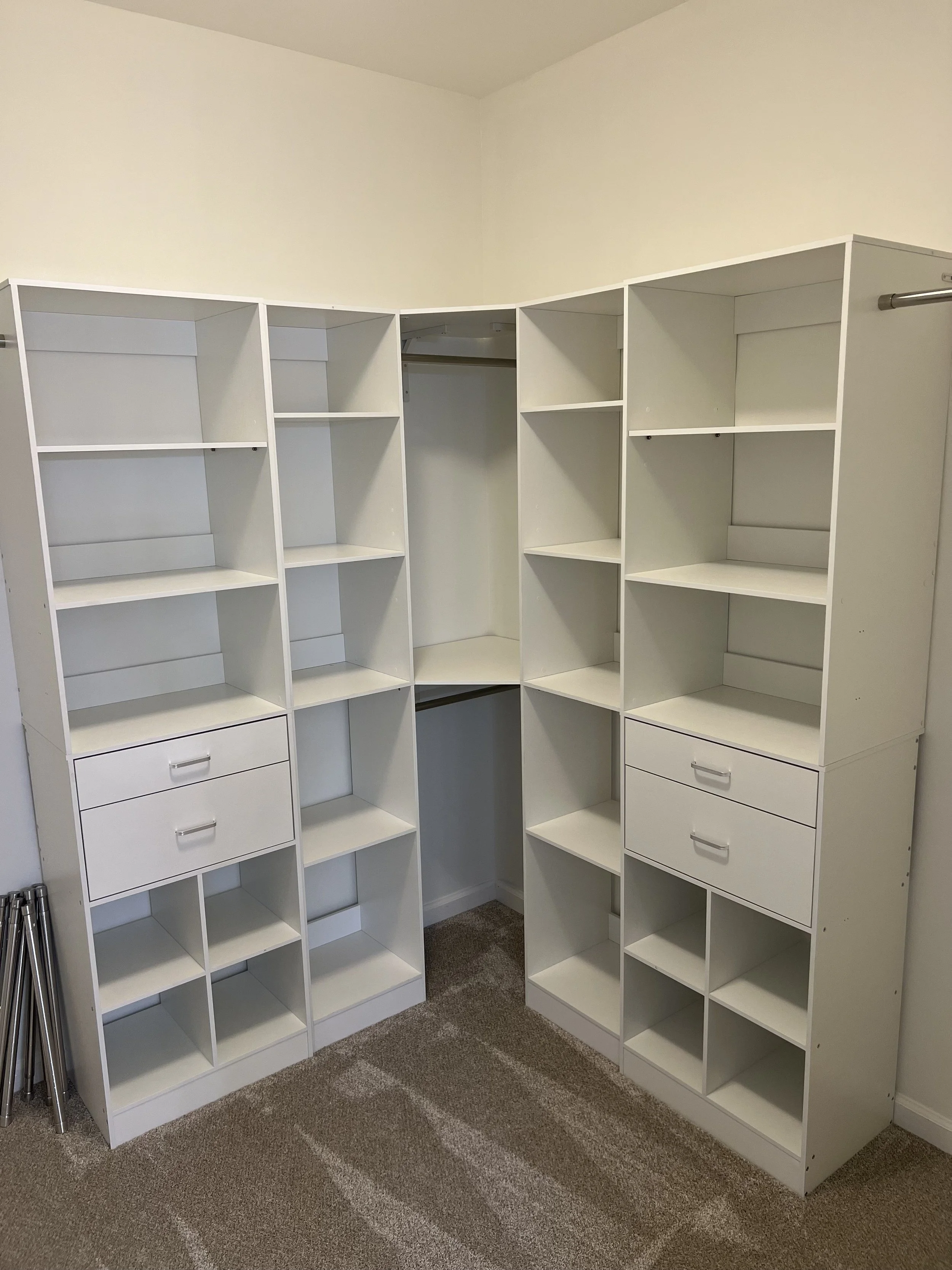 Empty white corner shelving unit with multiple compartments and drawers, placed in a room with beige carpet and white walls.