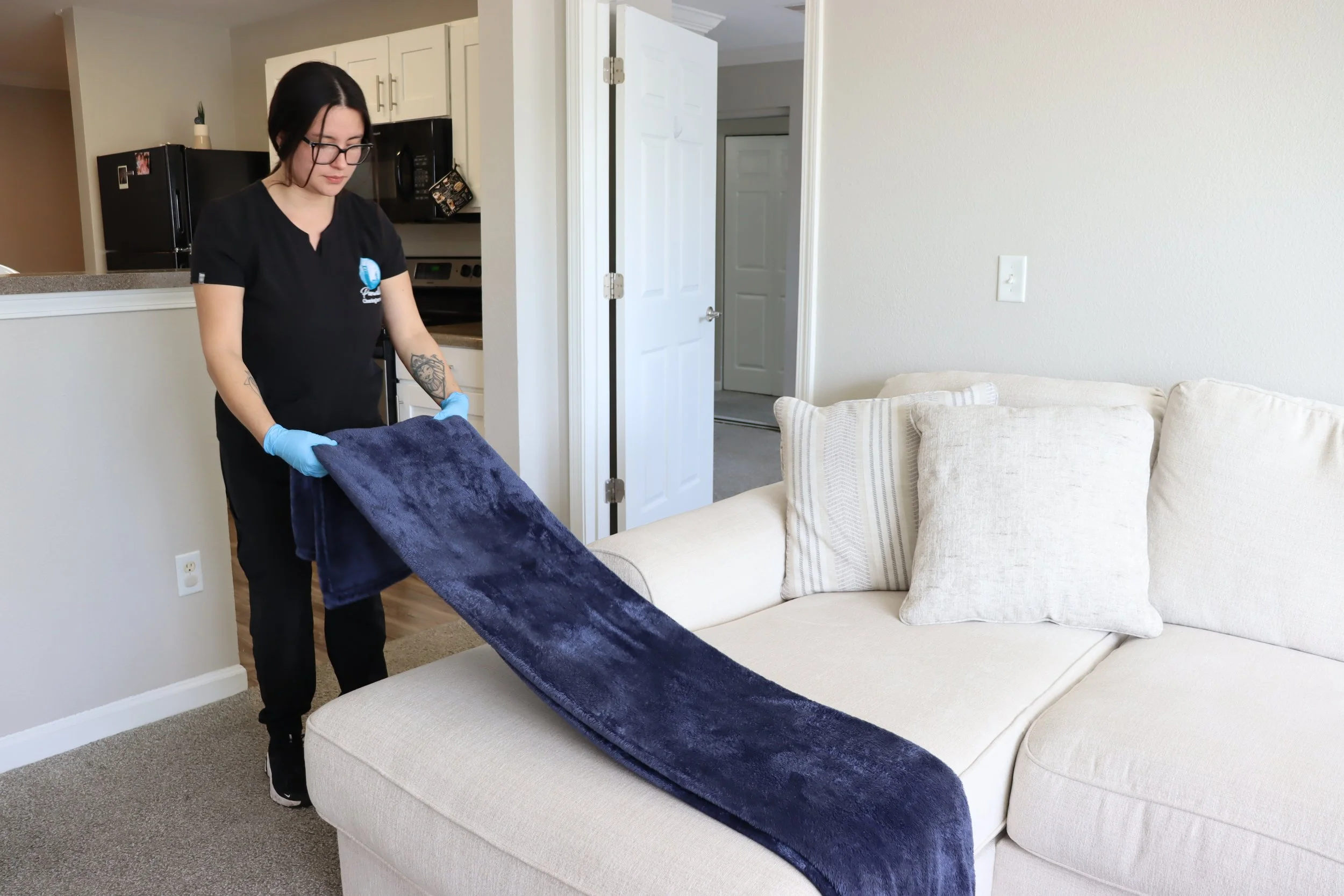 A Paradise Cleaning Team in black uniform, and blue gloves is vacuuming a beige sofa with a dark blue blanket draped over it in a living room.