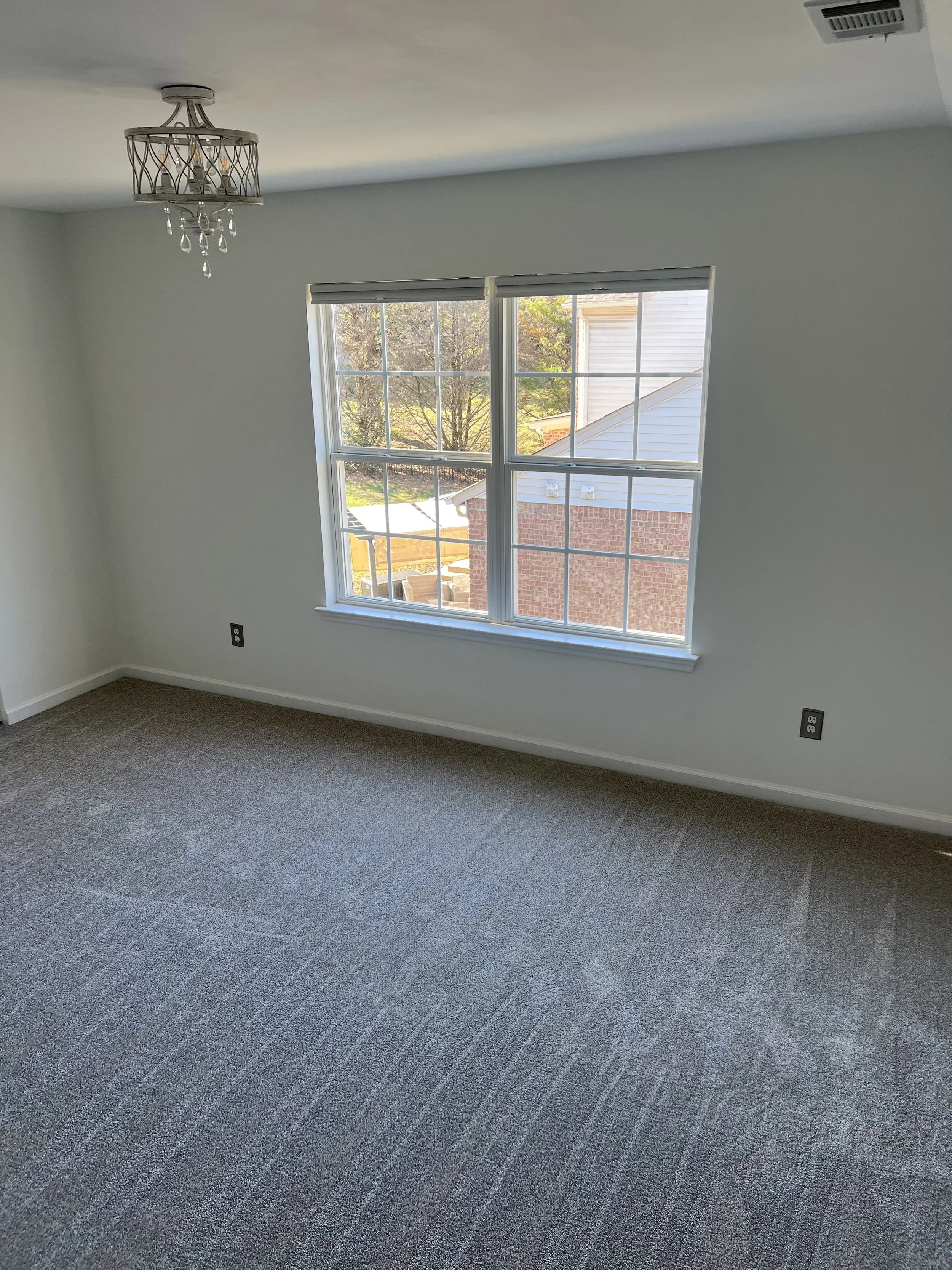 Empty room with carpet flooring, white walls, a large window with blinds, and a ceiling light fixture with decorative crystals.