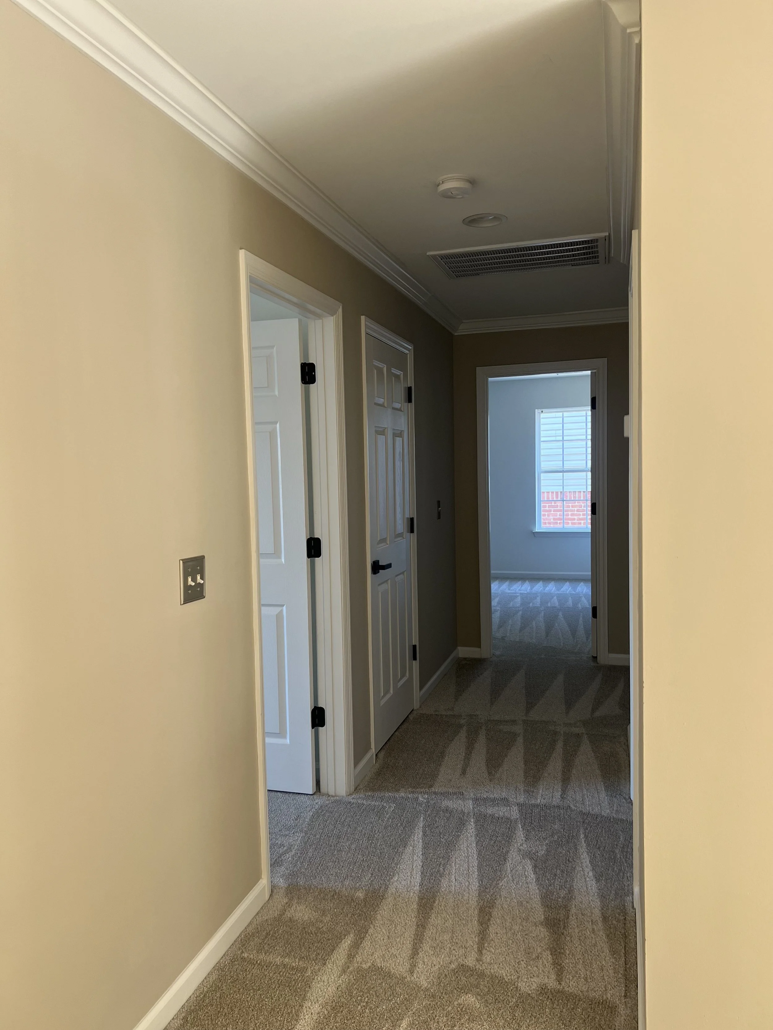 Hallway in a house with beige walls, white trim, carpeted floor, and two closed doors on the left, leading to other rooms, and a window at the end of the hall letting in natural light.