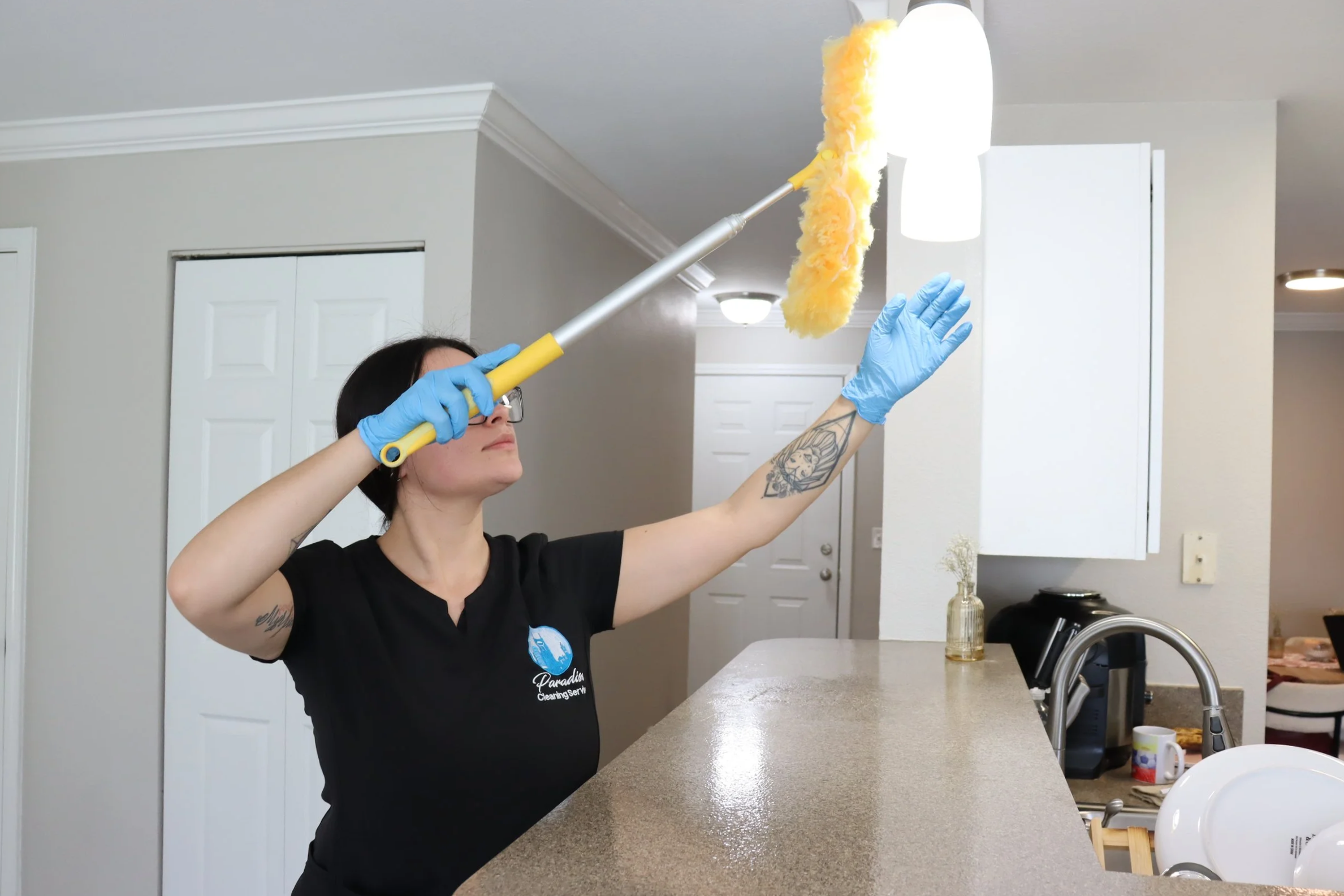 Cleaner dusting ceiling light fixture with extension duster during detailed home cleaning.