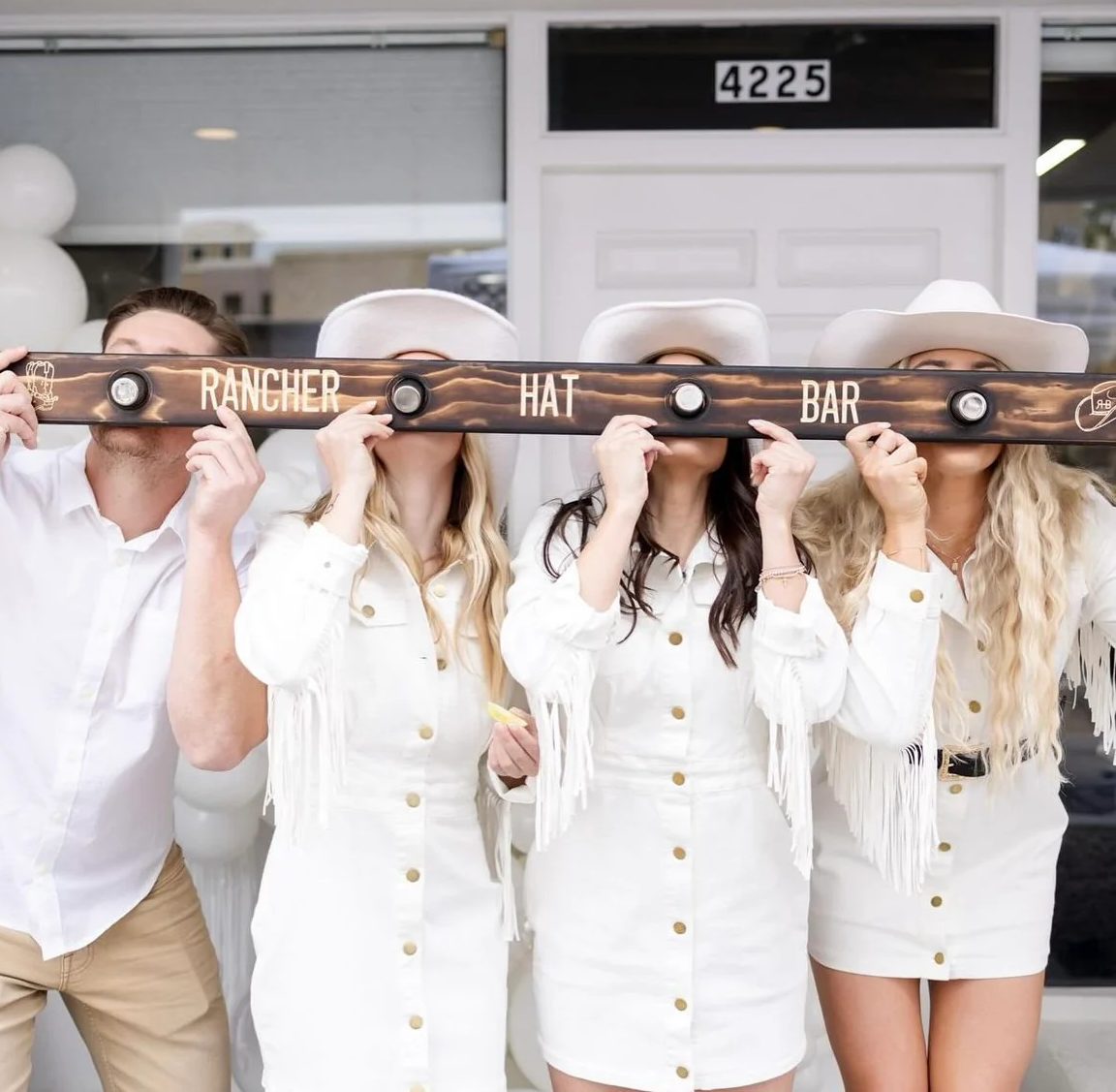 Four women dressed in white with white hats holding a bar with the words 'Rancher Hat Bar' in front of their faces. They stand in front of a building with the number 4225.