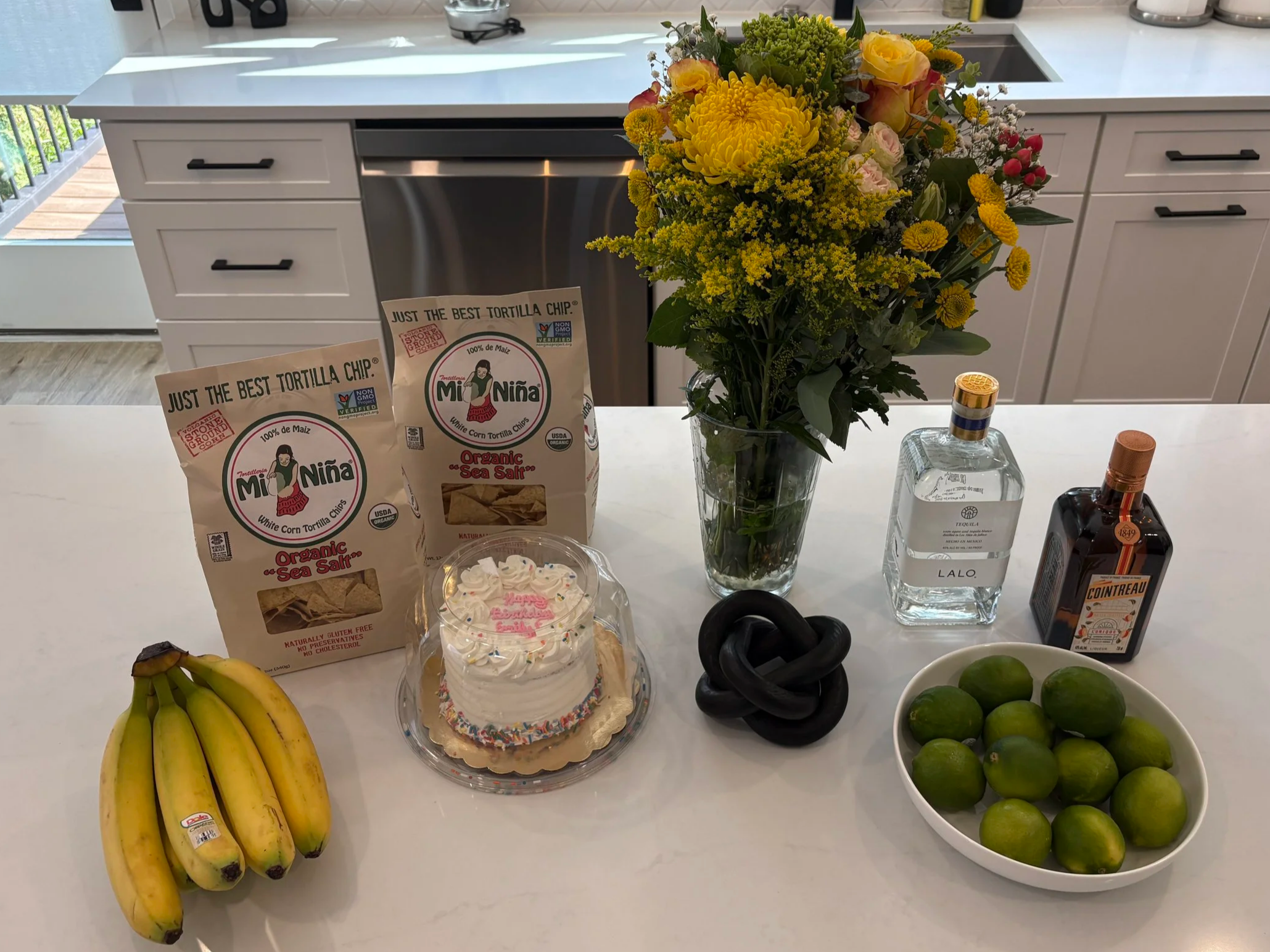 A kitchen counter with a bouquet of yellow, pink, and white flowers in a glass vase, bananas, limes in a bowl, two bags of tortilla chips, a cupcake with pink and white frosting, a bottle of tequila, a bottle of Cointreau, and a container of whipped cream.