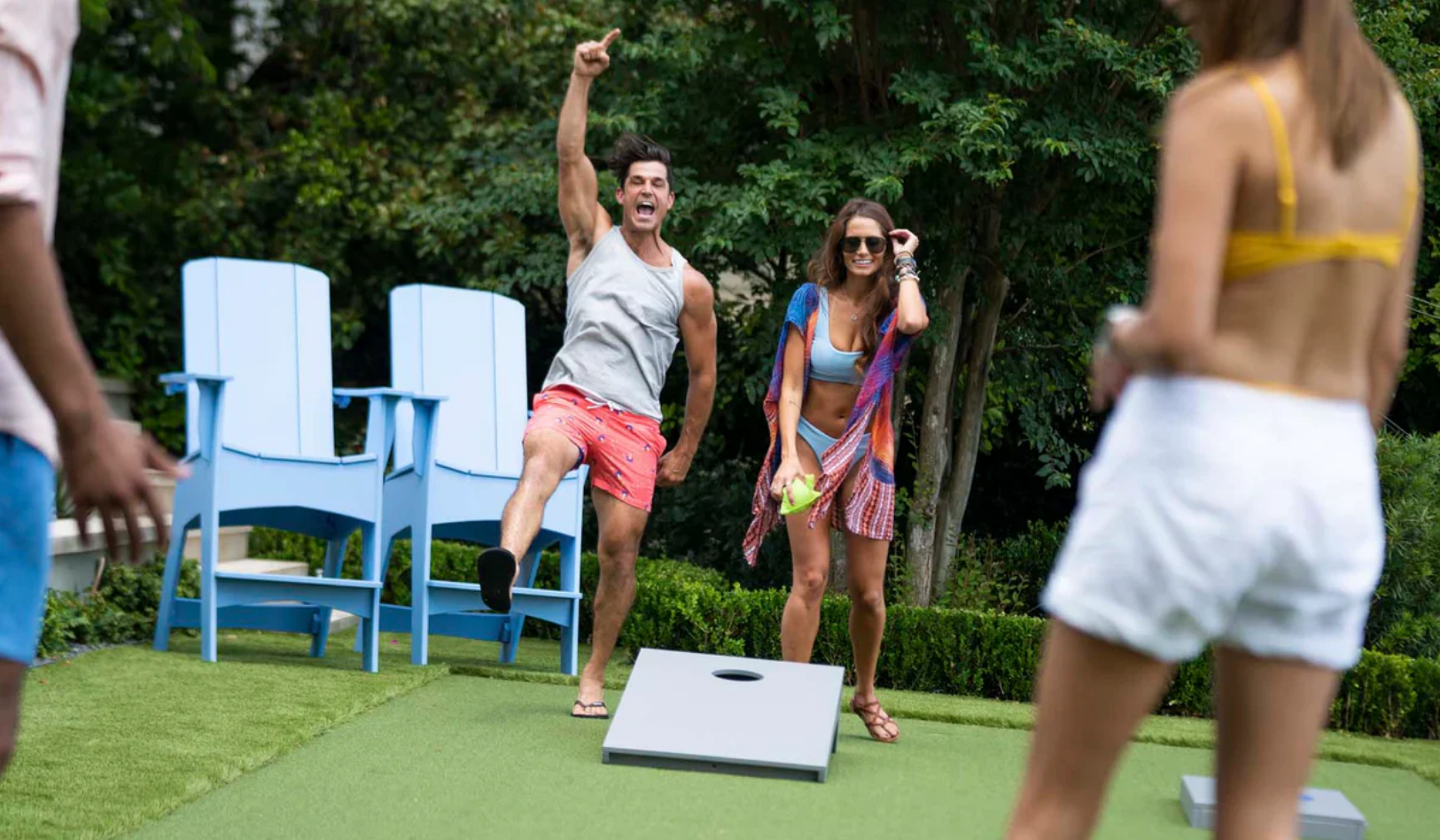 Group of friends playing cornhole outdoors on a sunny day, with trees and green grass in the background.