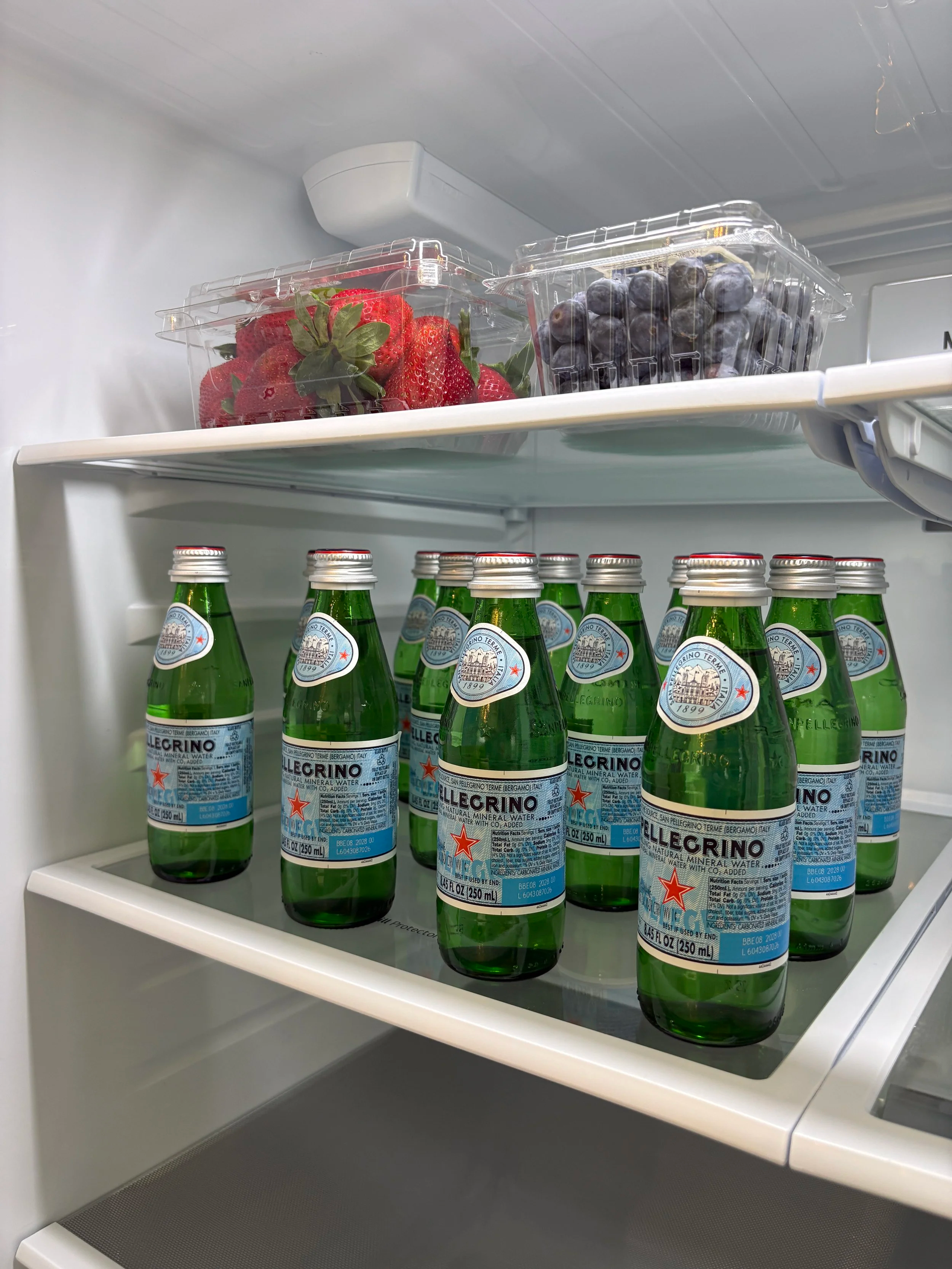 Open refrigerator shelf with several bottles of San Pellegrino sparkling water and containers of strawberries and blueberries.