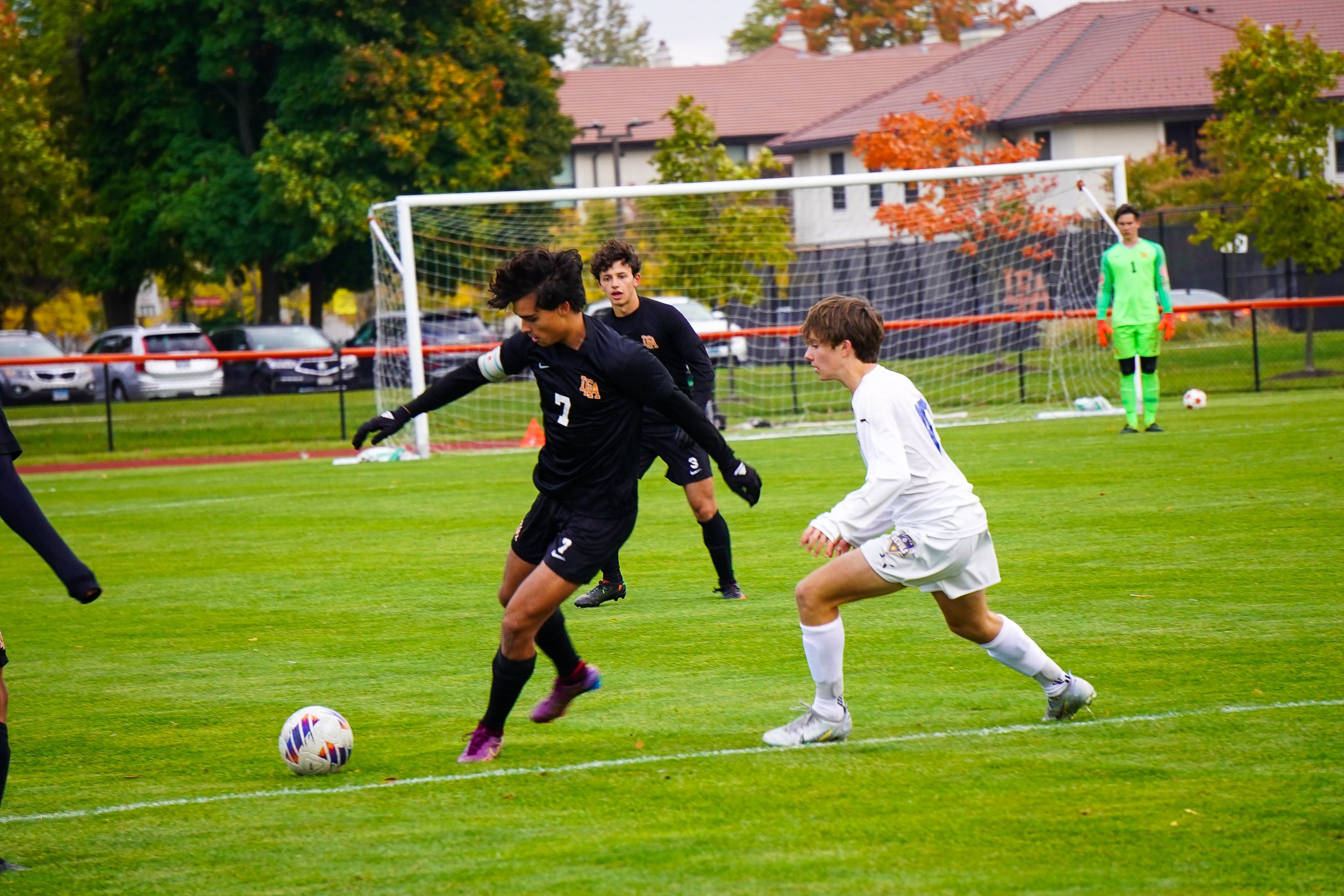 Soccer players competing for the ball during a match on a grass field, with a goalkeeper in a green uniform in the background.