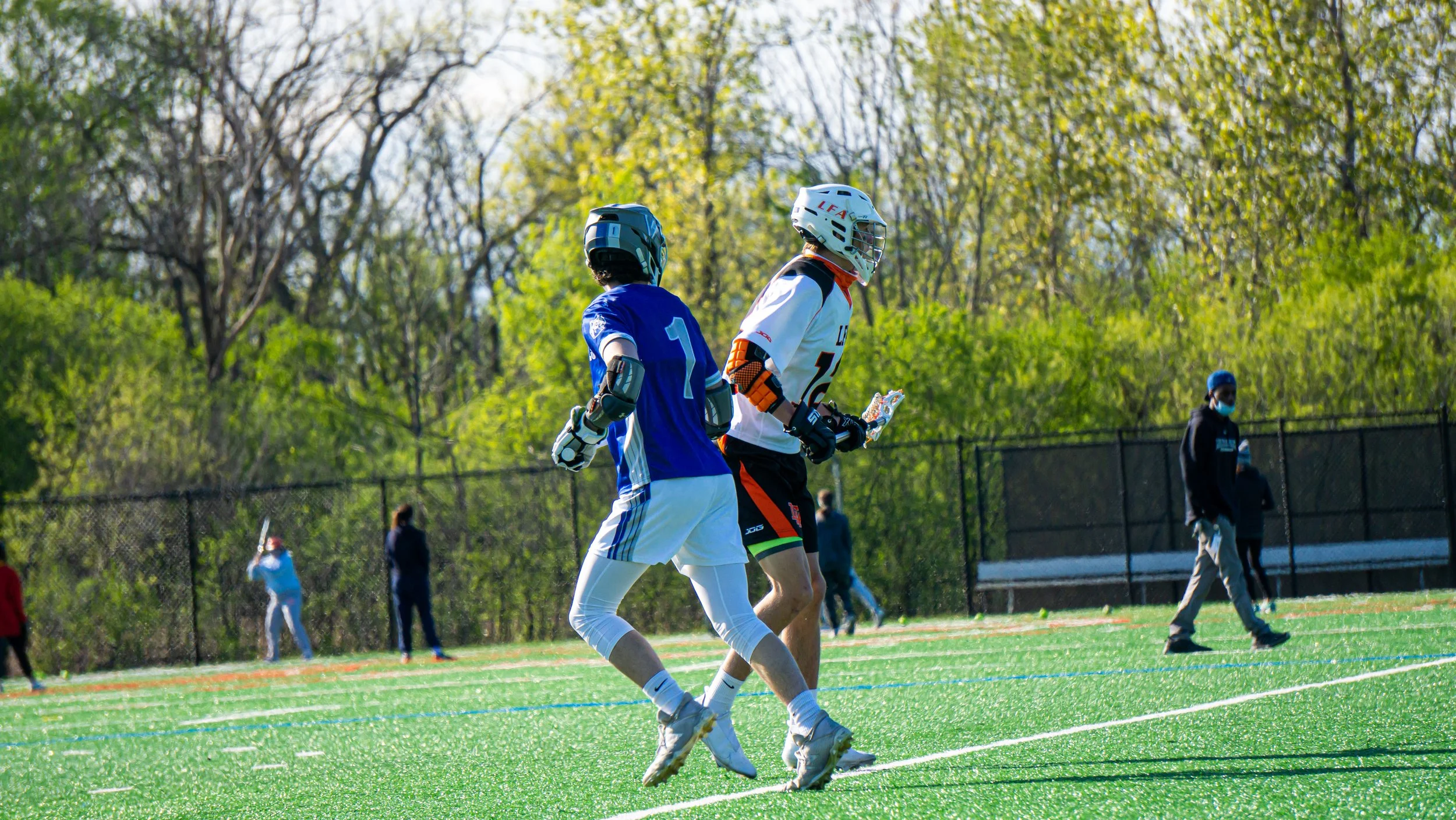 Two lacrosse players wearing helmets and protective gear, running on a green outdoor field with trees in the background.