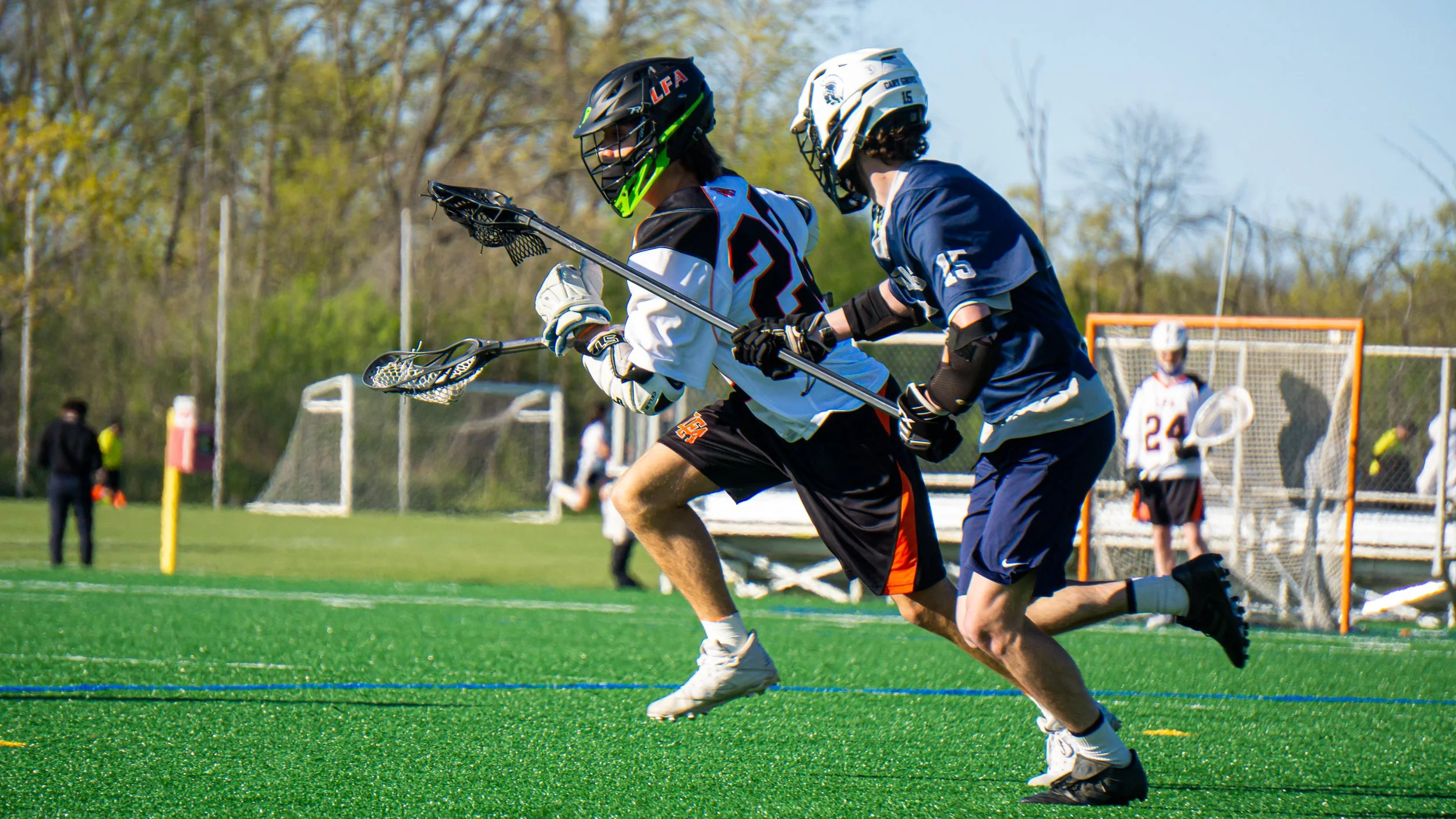 Two lacrosse players compete for possession of the ball on an outdoor field. One player wears a white and black jersey with the number 2, and the other wears a blue jersey with the number 15. Both players are wearing helmets, gloves, and protective g