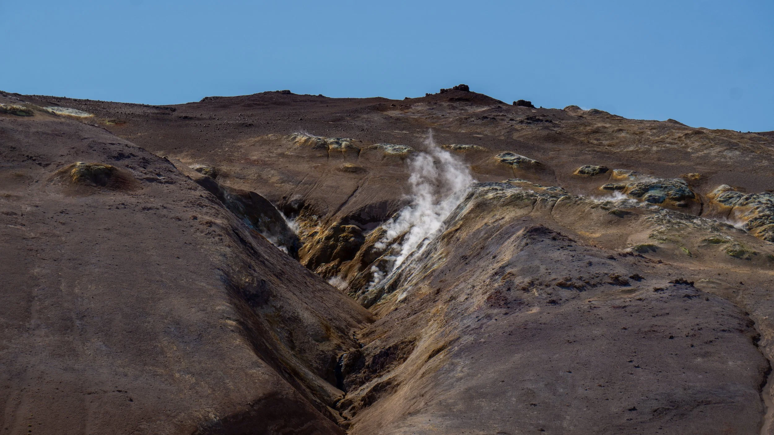 Steaming volcanic landscape with rugged, brown terrain and steaming vents against a clear blue sky.