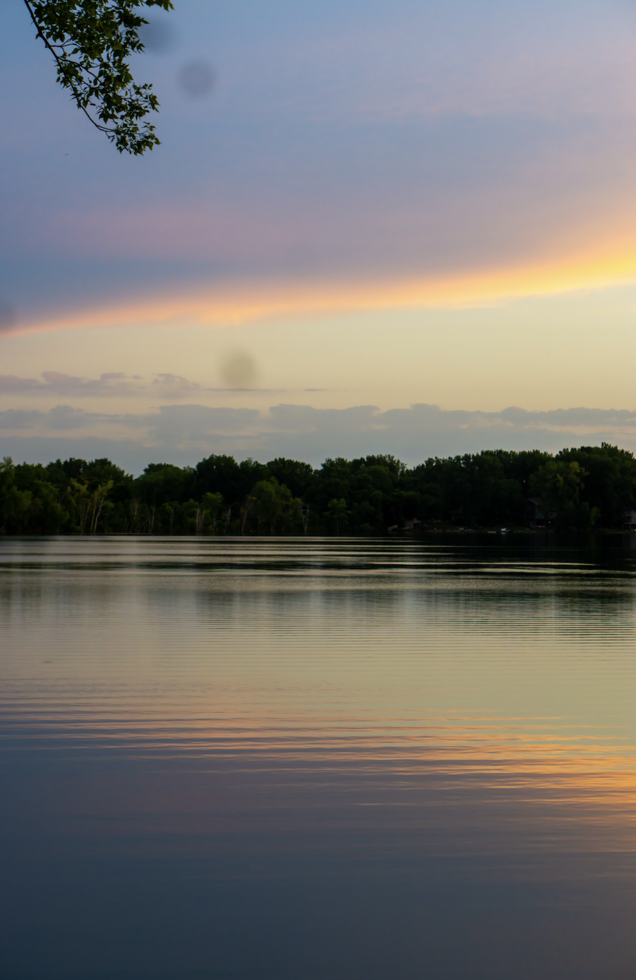 Calm lake at sunset with colorful sky and tree-lined shoreline in the distance.