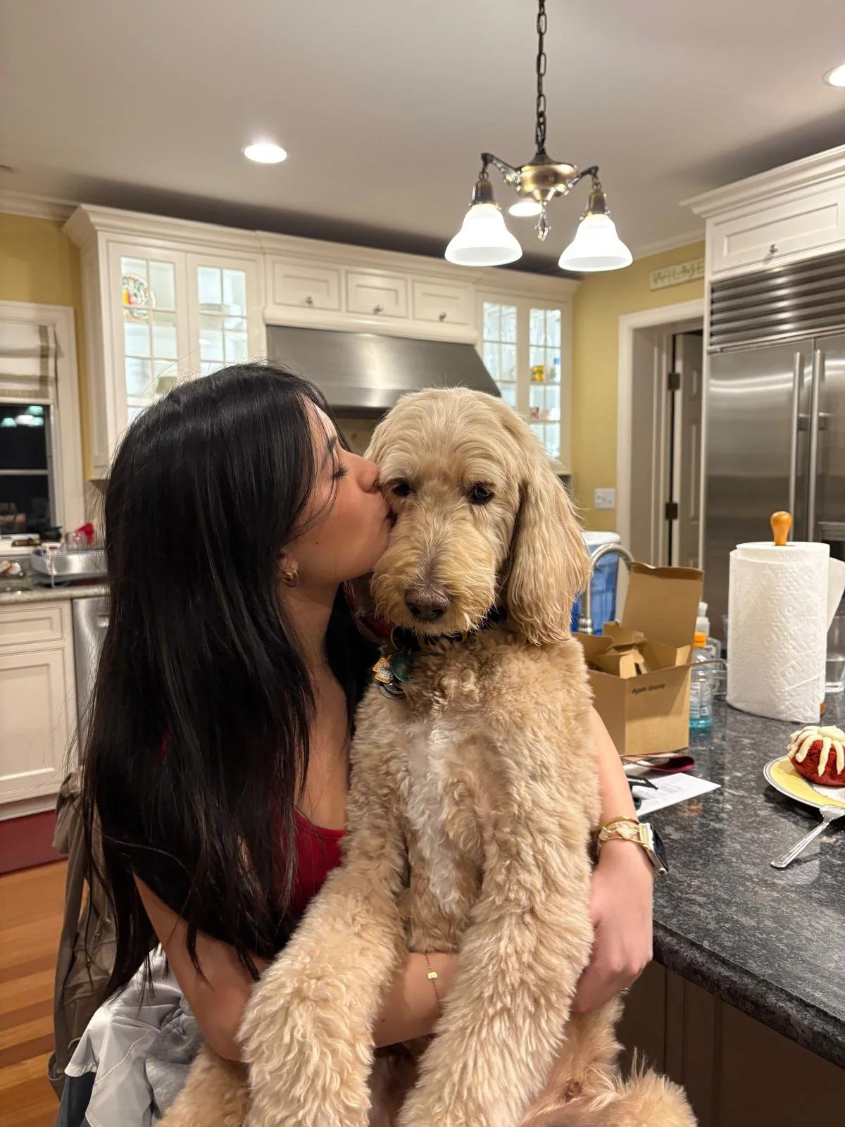 A woman with long dark hair is kissing a beige doodle-style dog on the face in a kitchen with white cabinets and stainless steel appliances.
