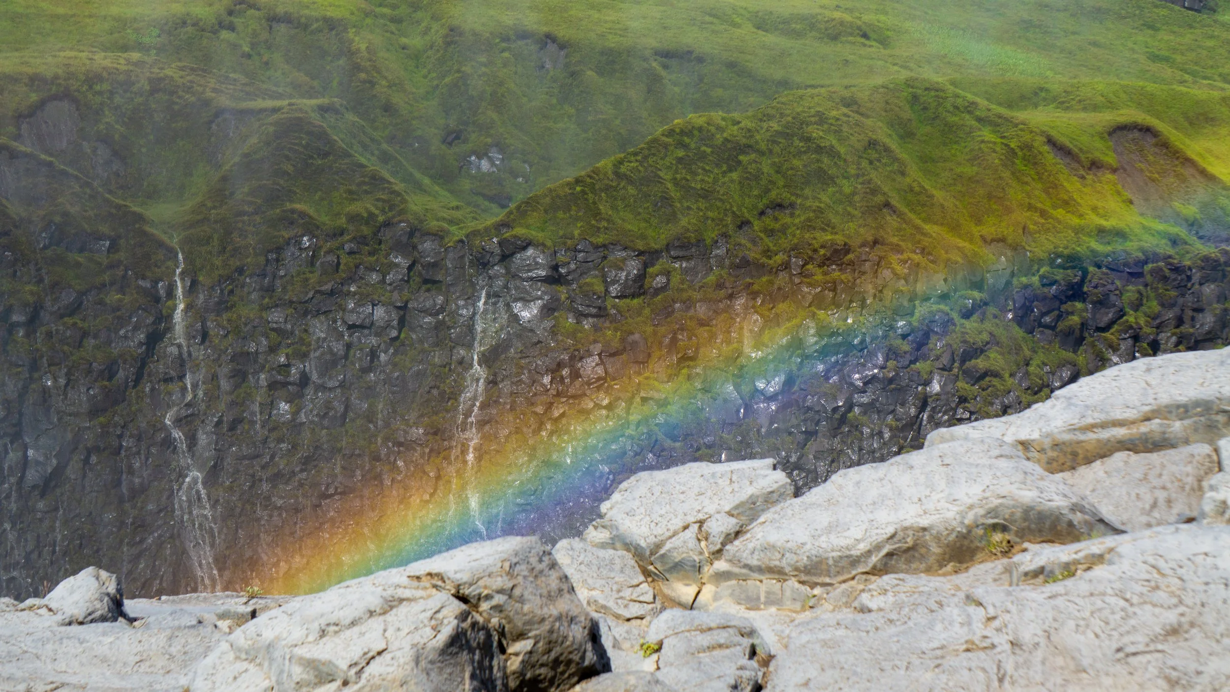 A vibrant rainbow arching across a rocky and mossy landscape with waterfalls and green hills in the background.