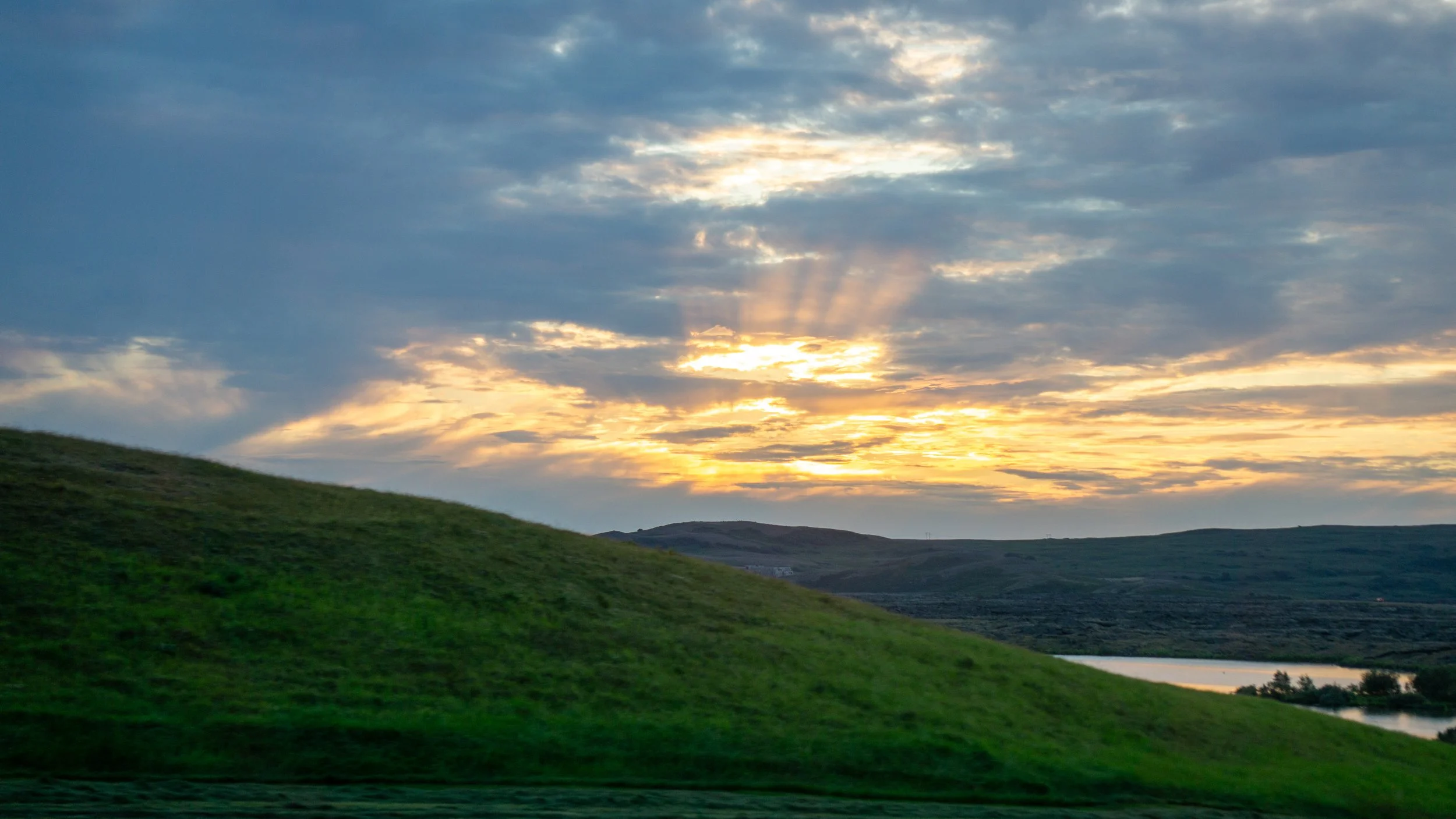 A landscape with a green hill, a body of water, and a sky with scattered clouds during sunset or sunrise.