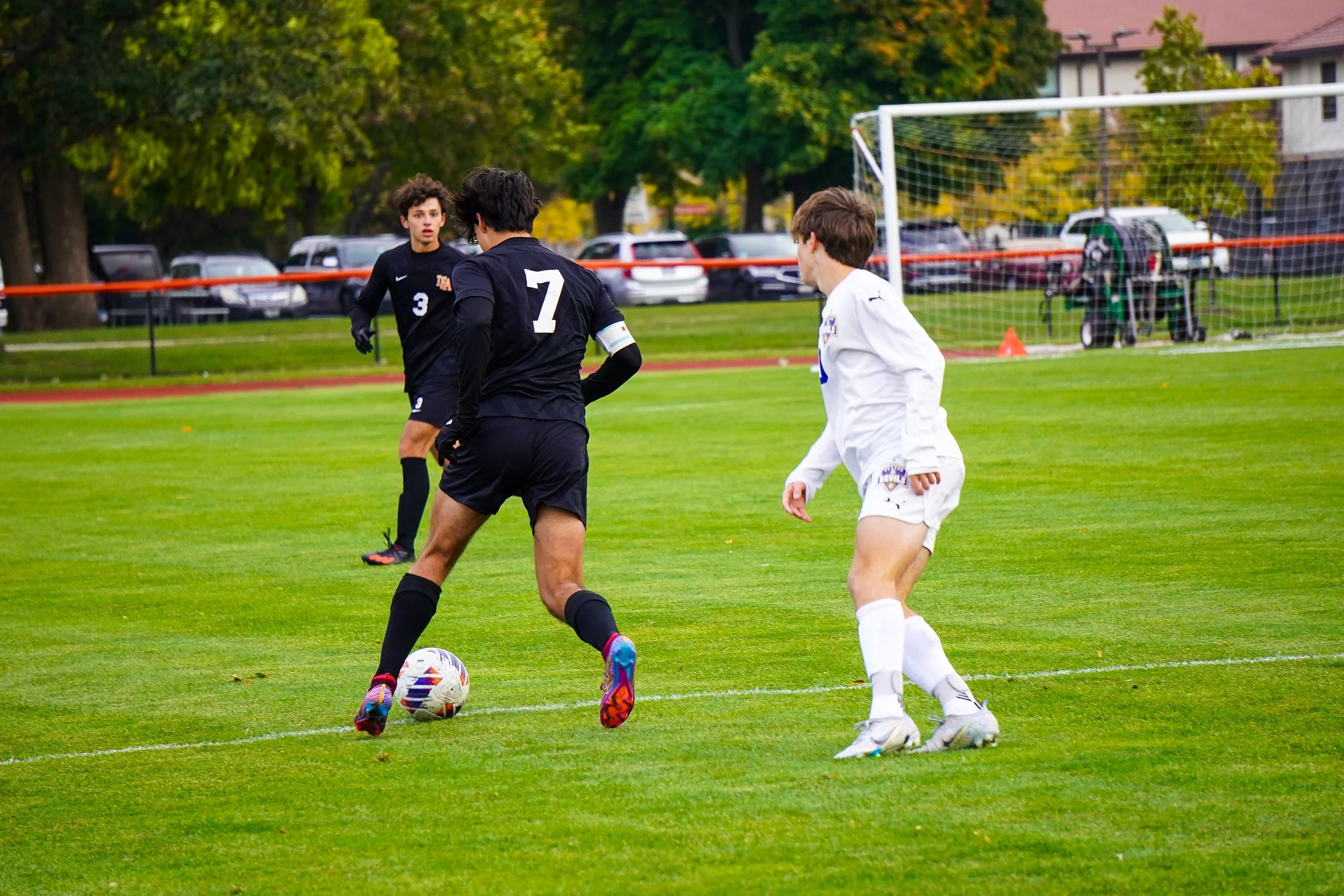 Soccer players in black and white uniforms on a green field during a game, with trees and parked cars in the background.
