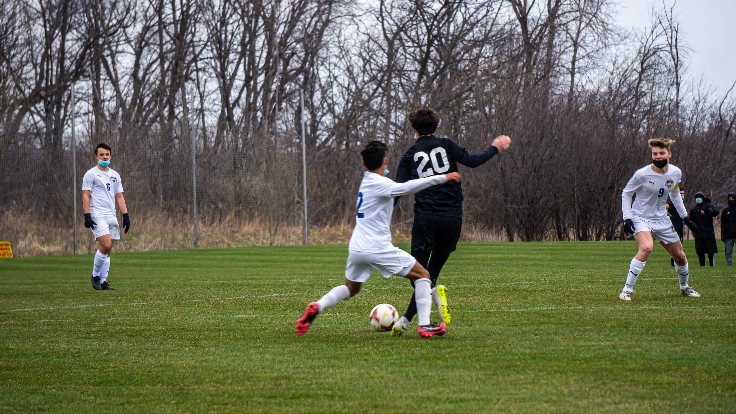 A soccer game taking place on a grassy field with four players visible. One player in a white jersey is kicking the ball, two others in white jerseys are nearby, and a player in a black jersey with the number 20 is defending. The background shows lea