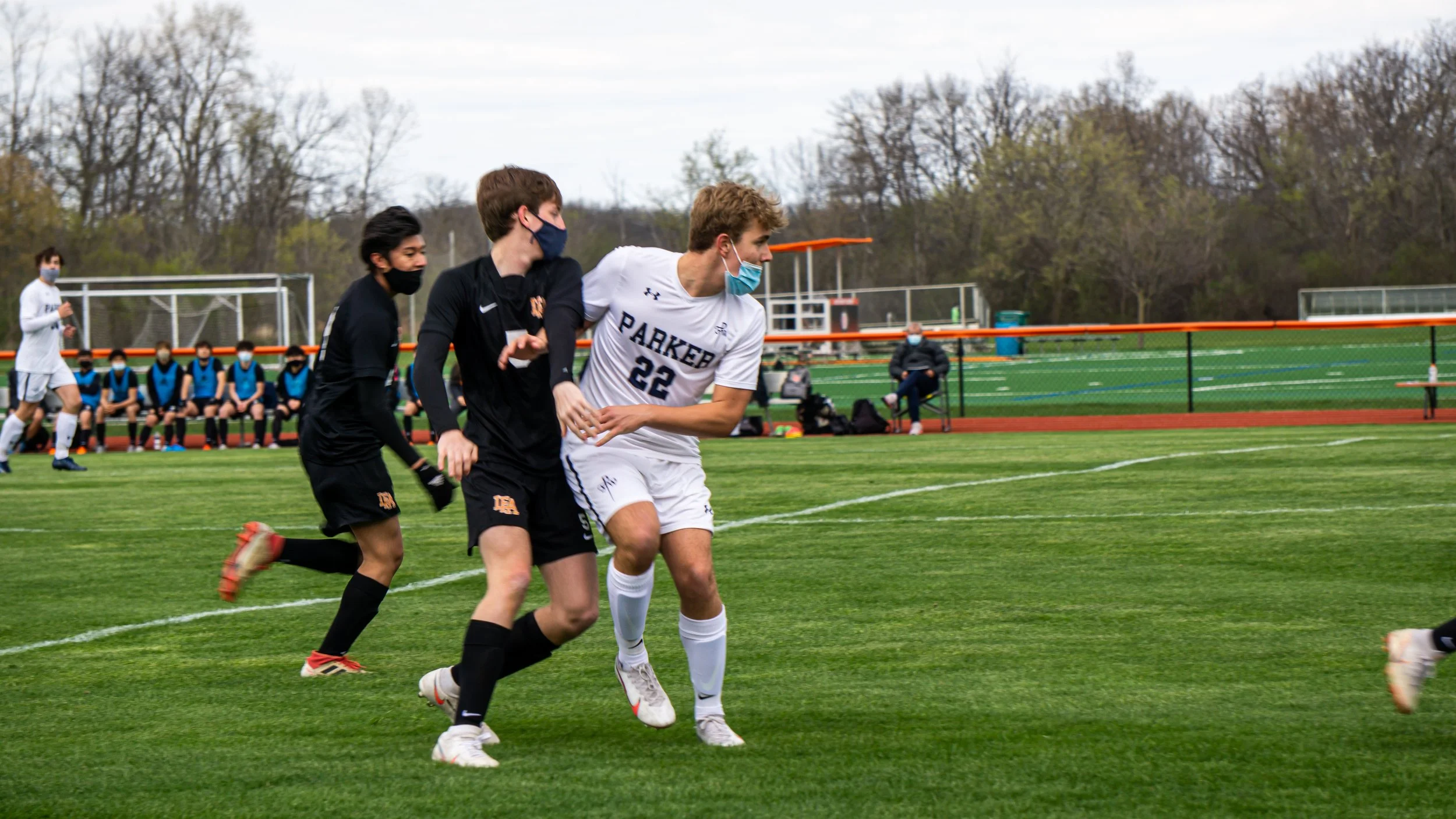 Soccer players wearing masks and school uniforms are playing on a green field with a school team sitting on the bench in the background.