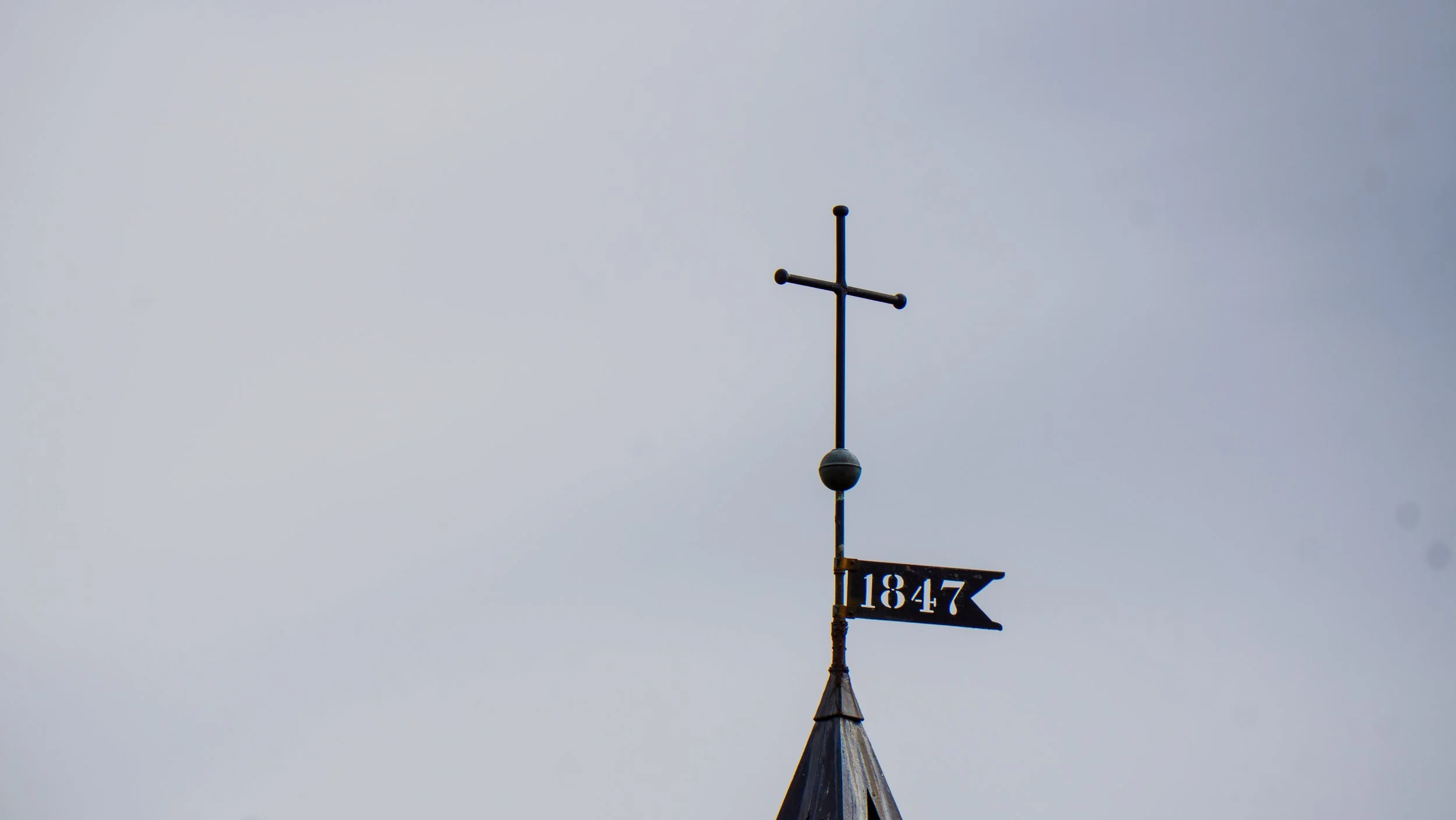 A weather vane with a cross at the top and a flag-shaped sign below displaying the year 1847, mounted on top of a building against a cloudy sky.