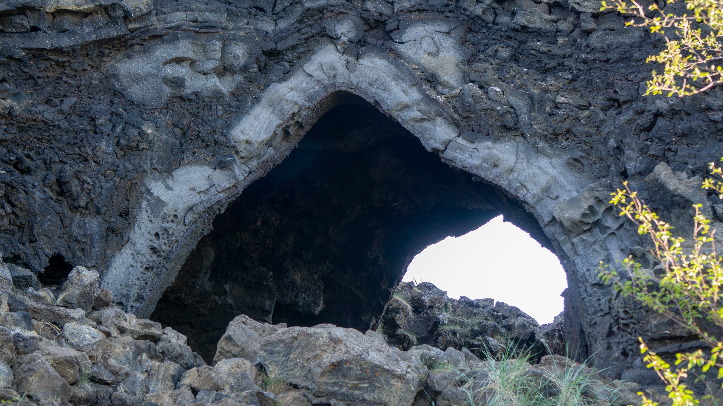 A natural arch formation in a rocky landscape with dark volcanic rock, small green plants, and bright sky visible through the opening.