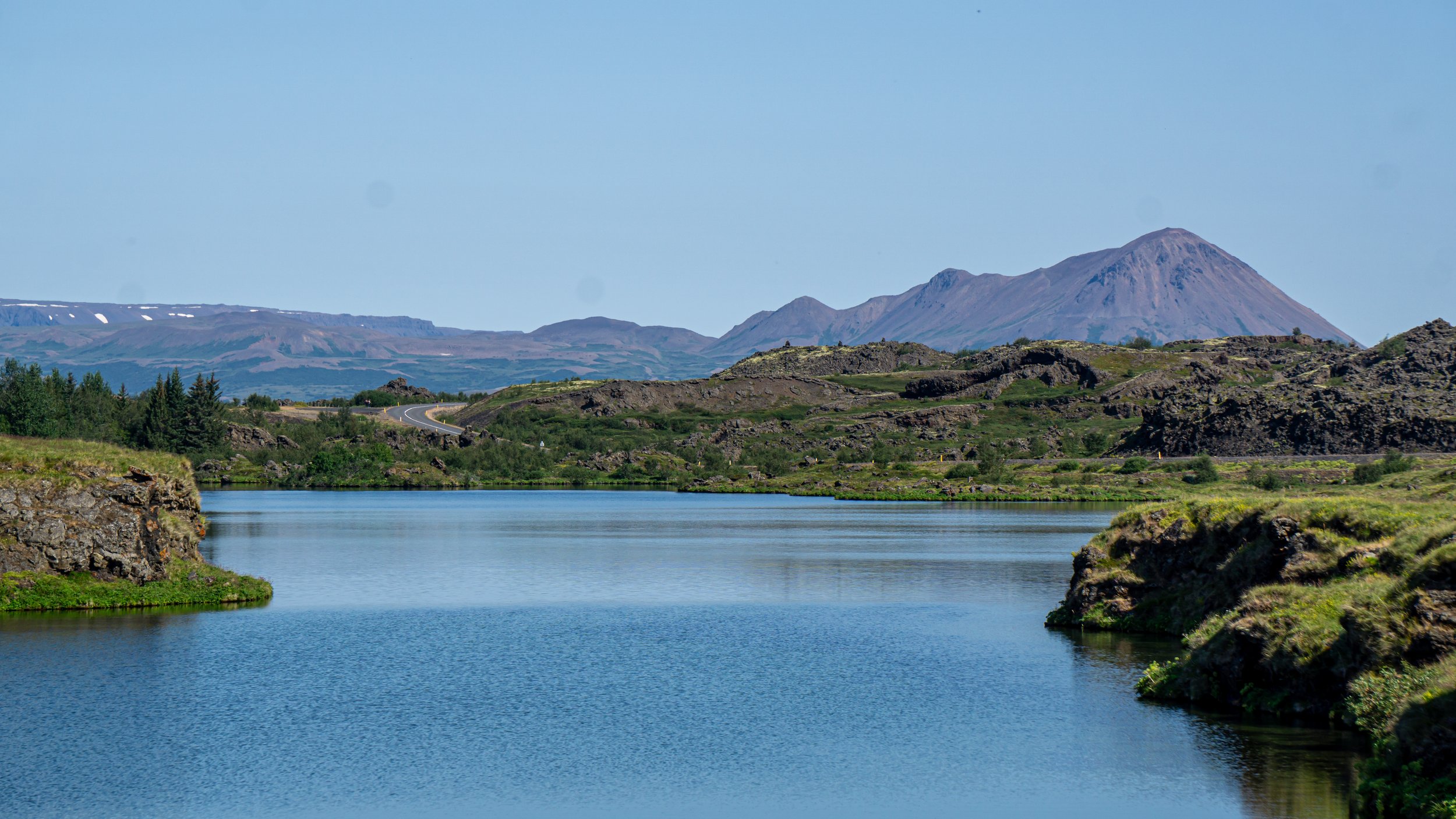 A serene landscape featuring a calm river with green, rocky banks, winding through a wide valley. In the background, there are rugged hills and distant mountains with some snow patches, under a clear blue sky.