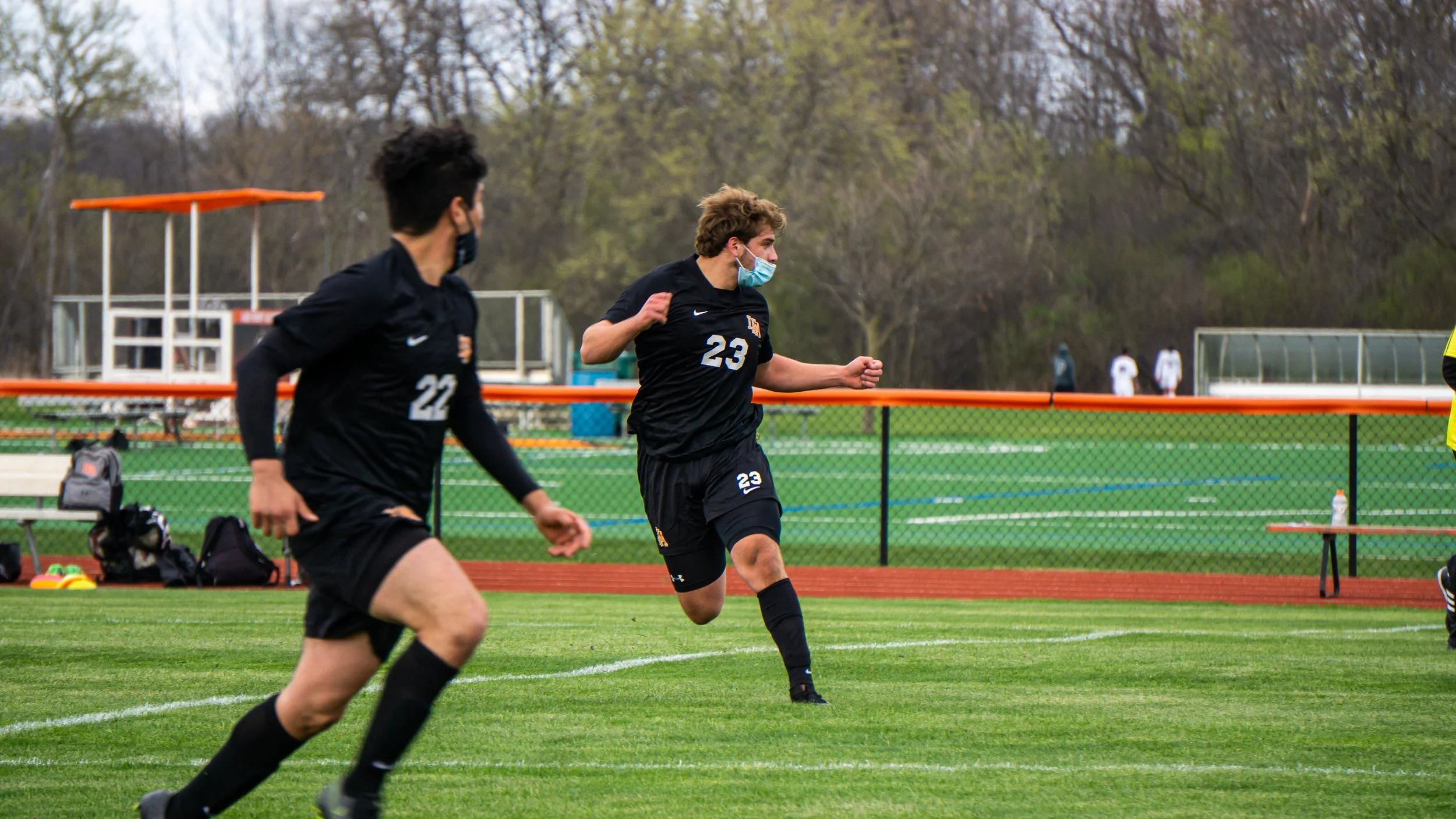 Two soccer players wearing black uniforms and face masks running on the field during a game, with a fence, coach's benches, and trees in the background.