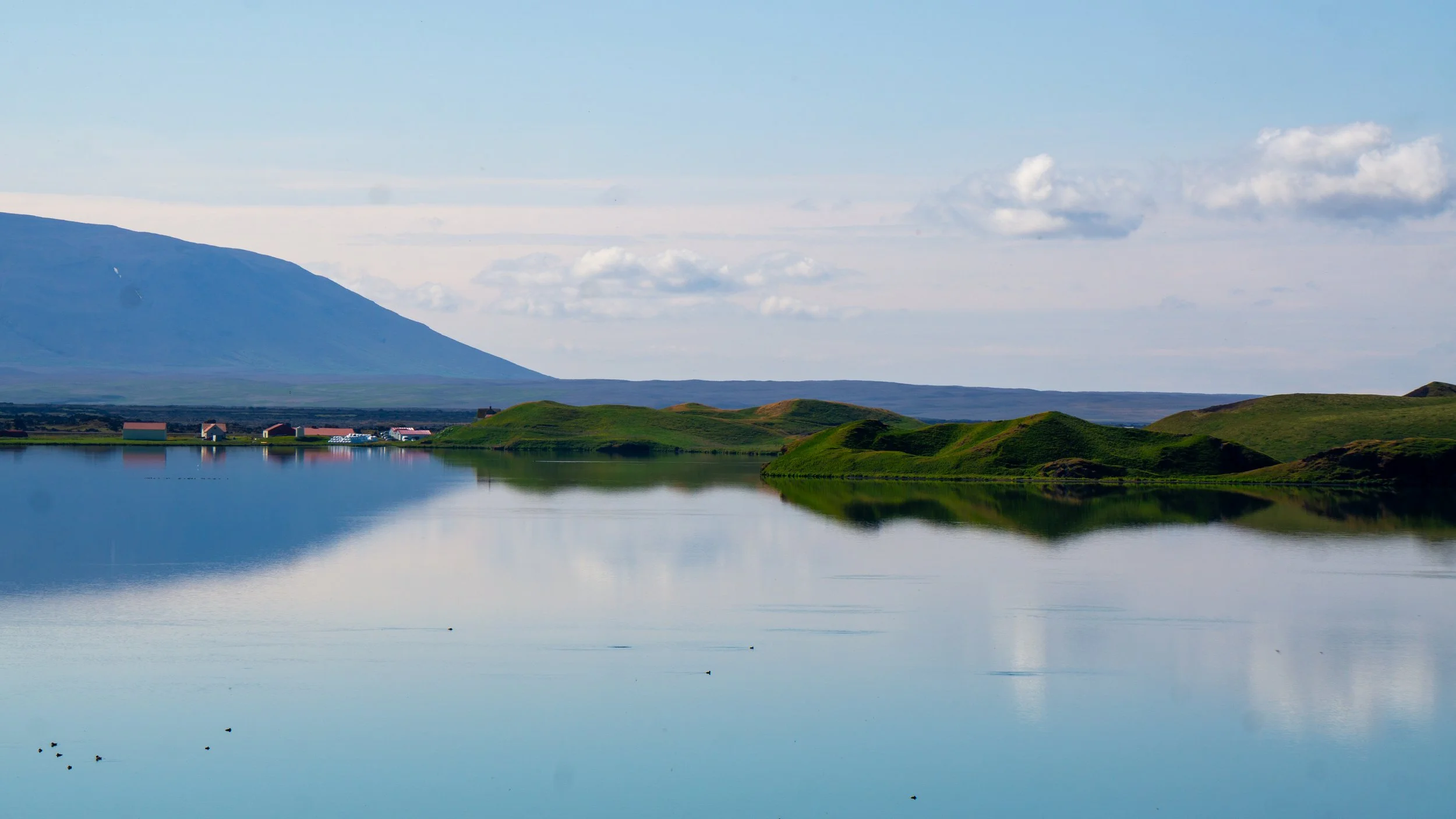 A calm lake reflecting green hills and scattered buildings, with a mountain in the background under a partly cloudy sky.