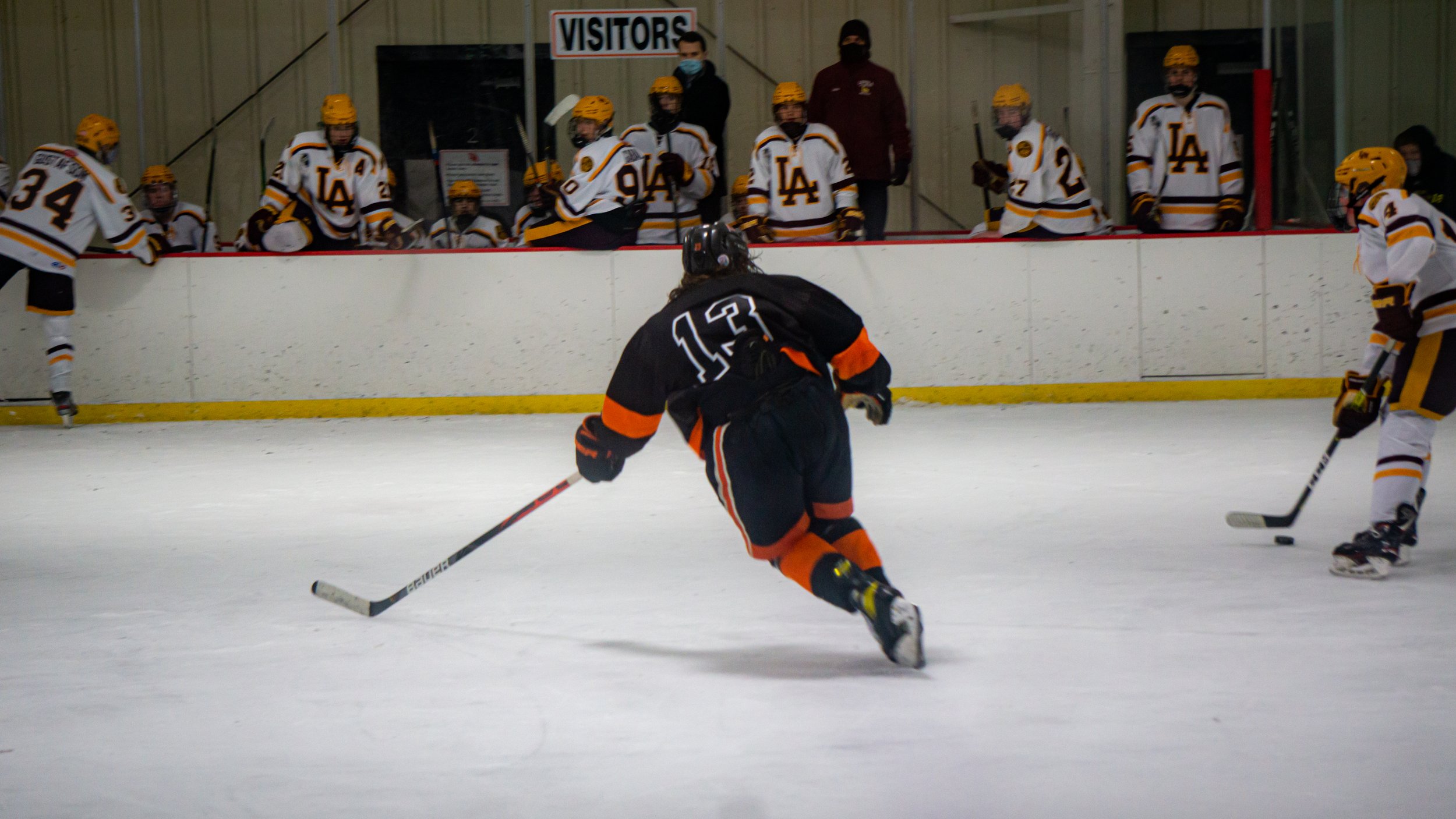 Hockey player in black jersey shoots puck on ice rink with team in white jerseys watching from bench